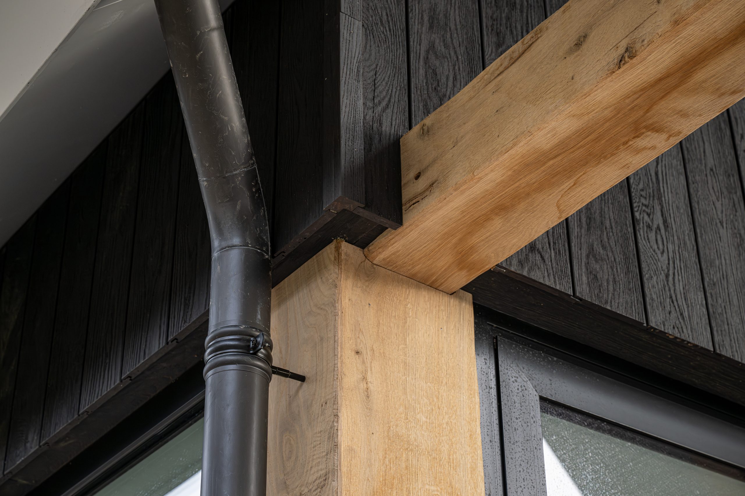 Close-up van de nauwkeurige afwerking van eikenhouten balkbekleding bij de aansluiting met een zwarte regenpijp en gevel.