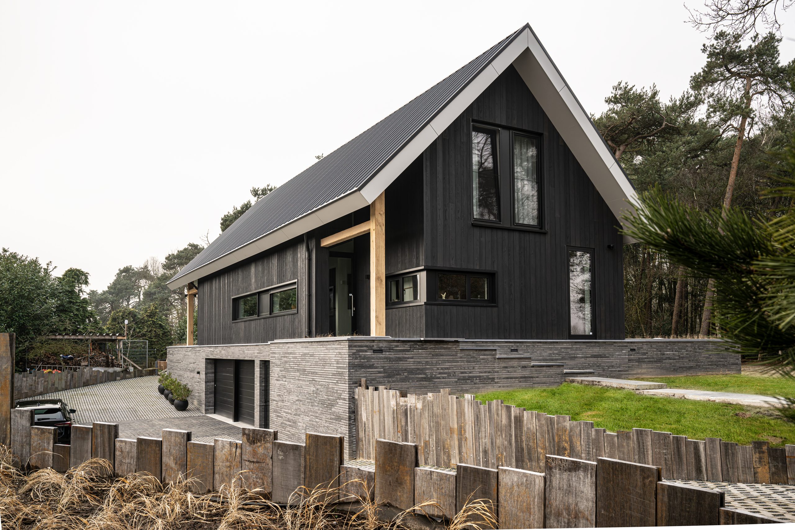 Zijaanzicht van de woning met eikenhouten kolommen, gezien door de takken van een dennenboom voor een natuurlijk effect.