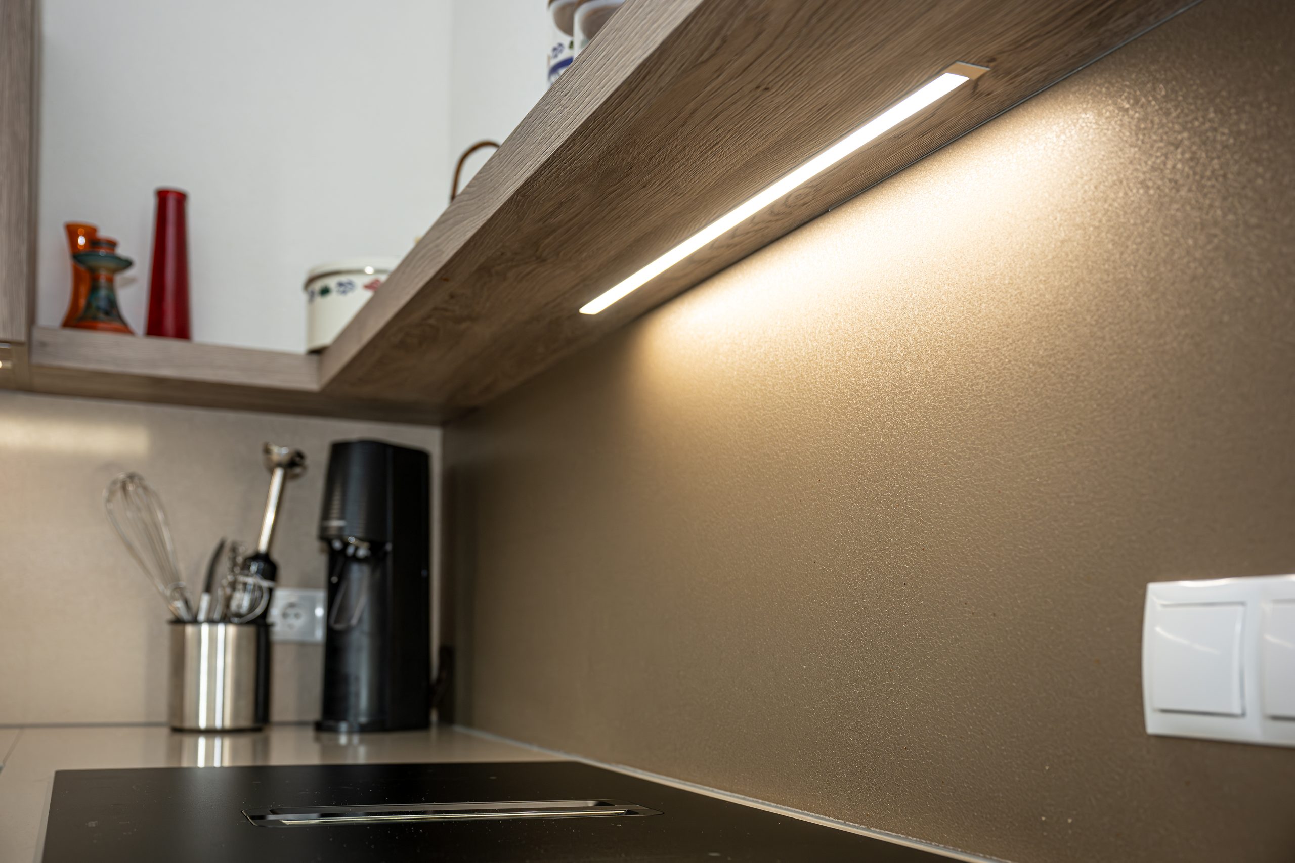 Detailopname van de geïntegreerde LED-verlichting onder een houten keukenplank, die schijnt op de getextureerde achterwand en keukengerei.