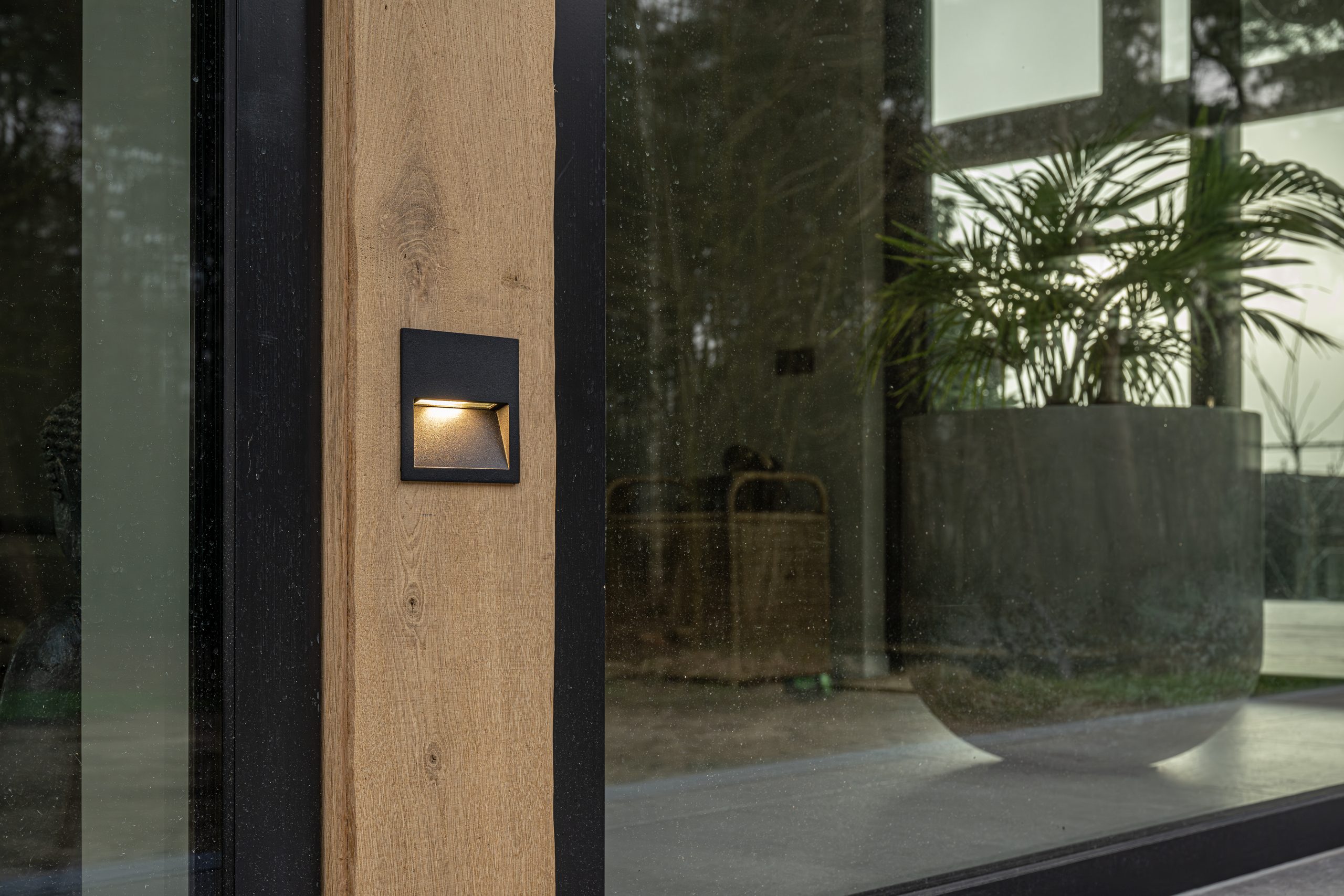 Detailopname van een eikenhouten balkomtimmering met een ingebouwde zwarte vierkante wandlamp voor sfeervolle buitenverlichting.