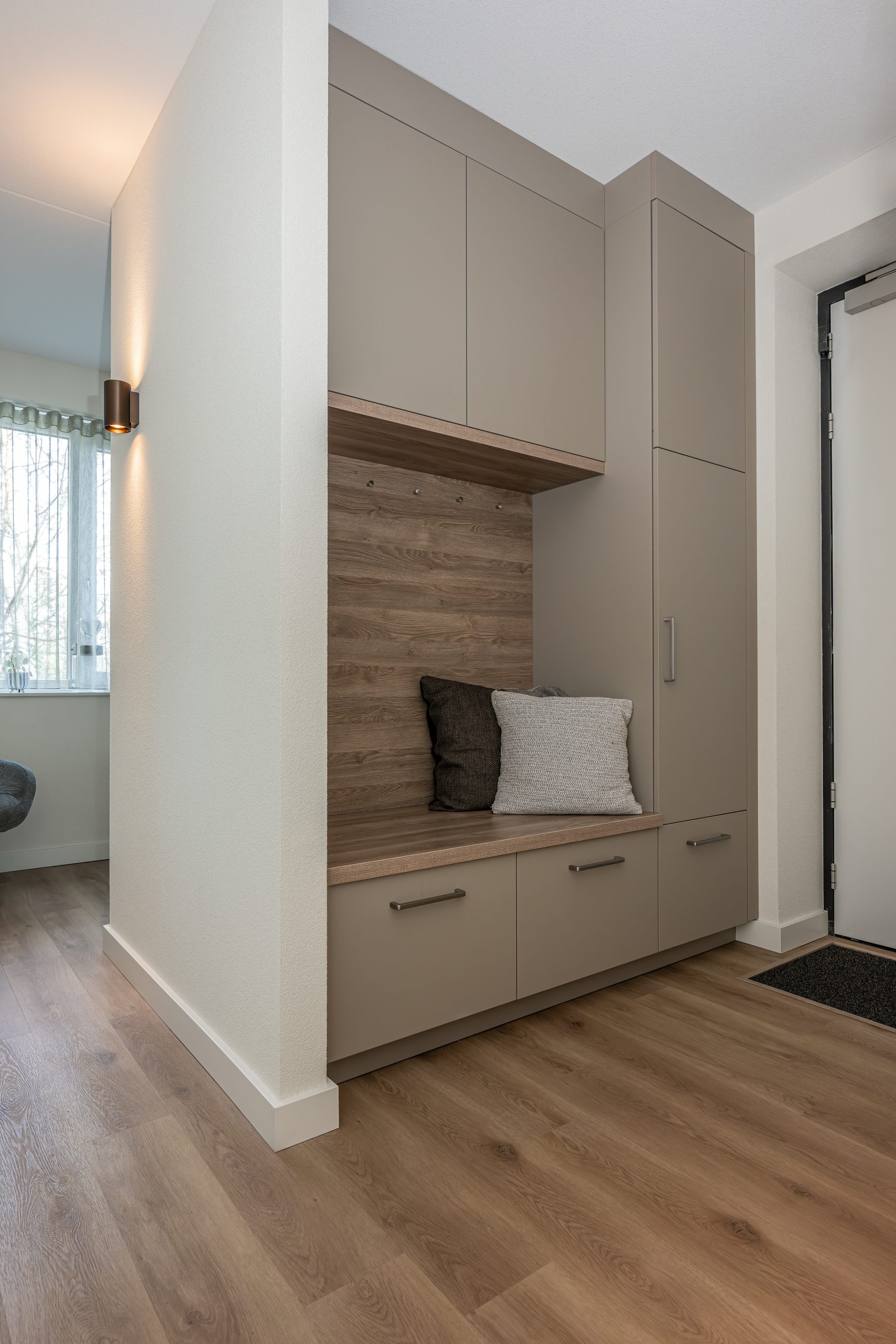 Een op maat gemaakt halmeubel met grijze inbouwkasten en een nis met een houten zitbankje, kussens en kapstokhaken.