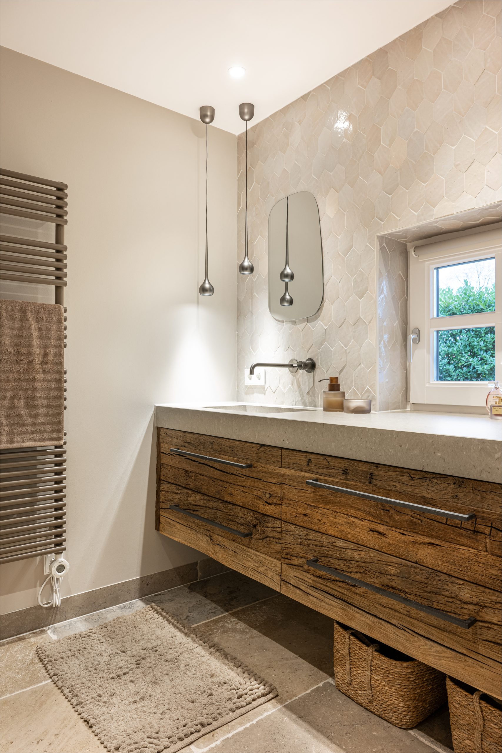 Robuust houten badkamermeubel met betonlook blad, gecombineerd met beige hexagon wandtegels en sfeervolle hanglampen.