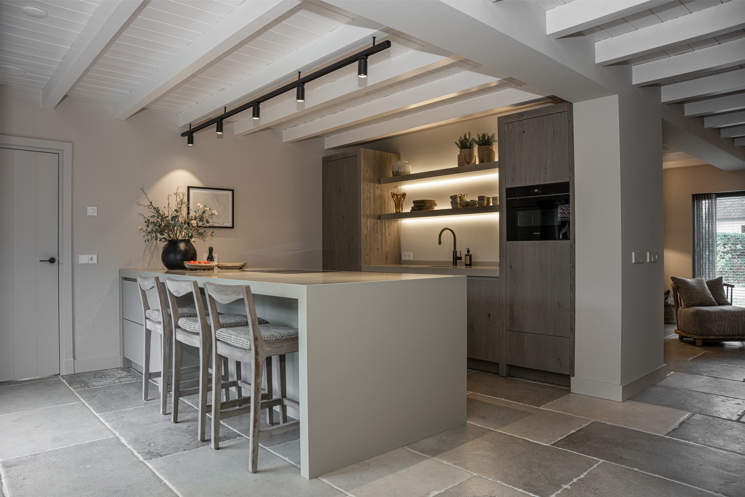 Moderne landelijke keuken met een groot kookeiland, houten barkrukken, inbouwverlichting en een grijze natuurstenen vloer.