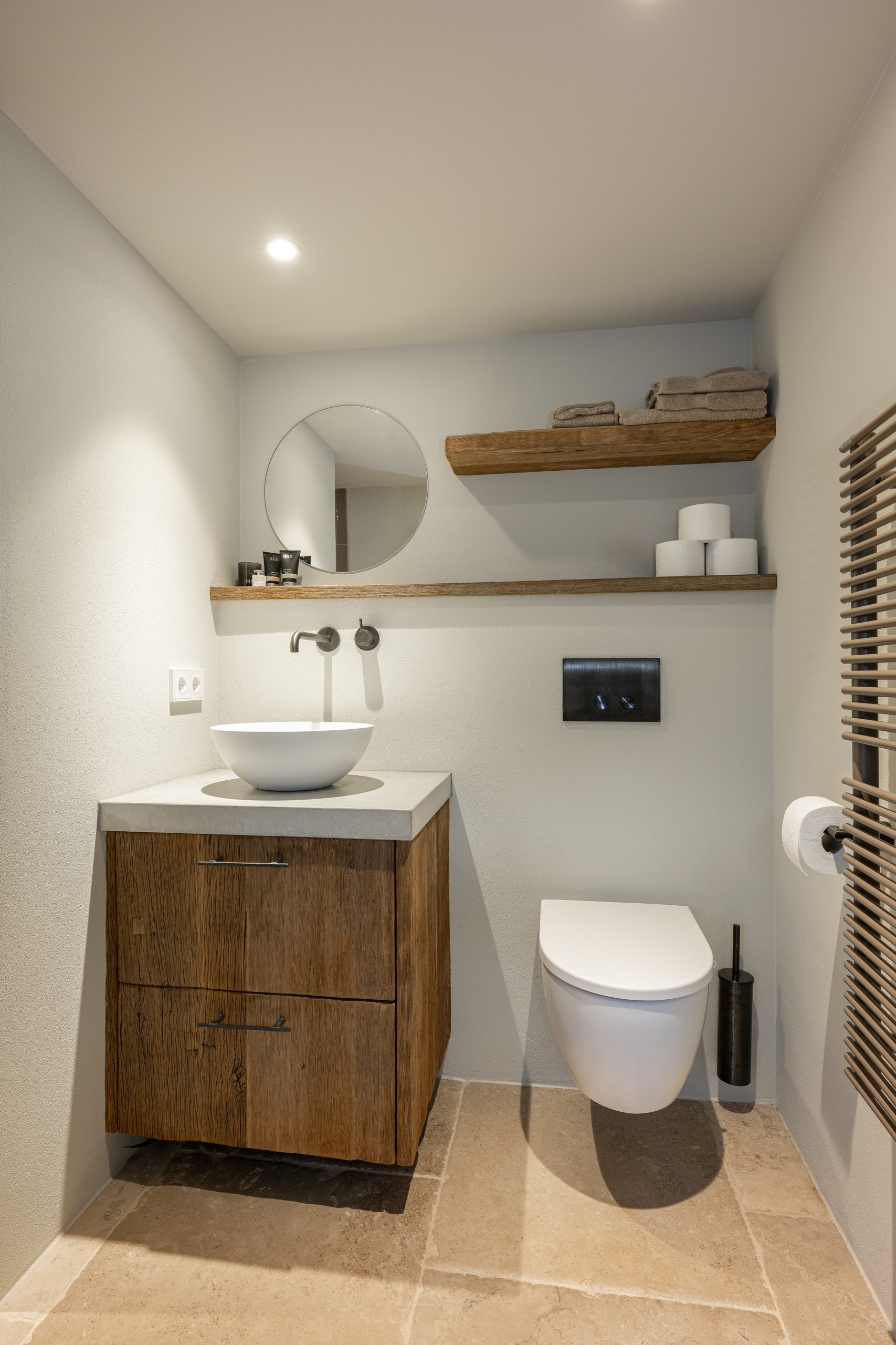 Stijlvolle toiletruimte voorzien van een houten wastafelmeubel met witte opzetwaskom, bijpassende wandplanken en een design handdoekradiator.
