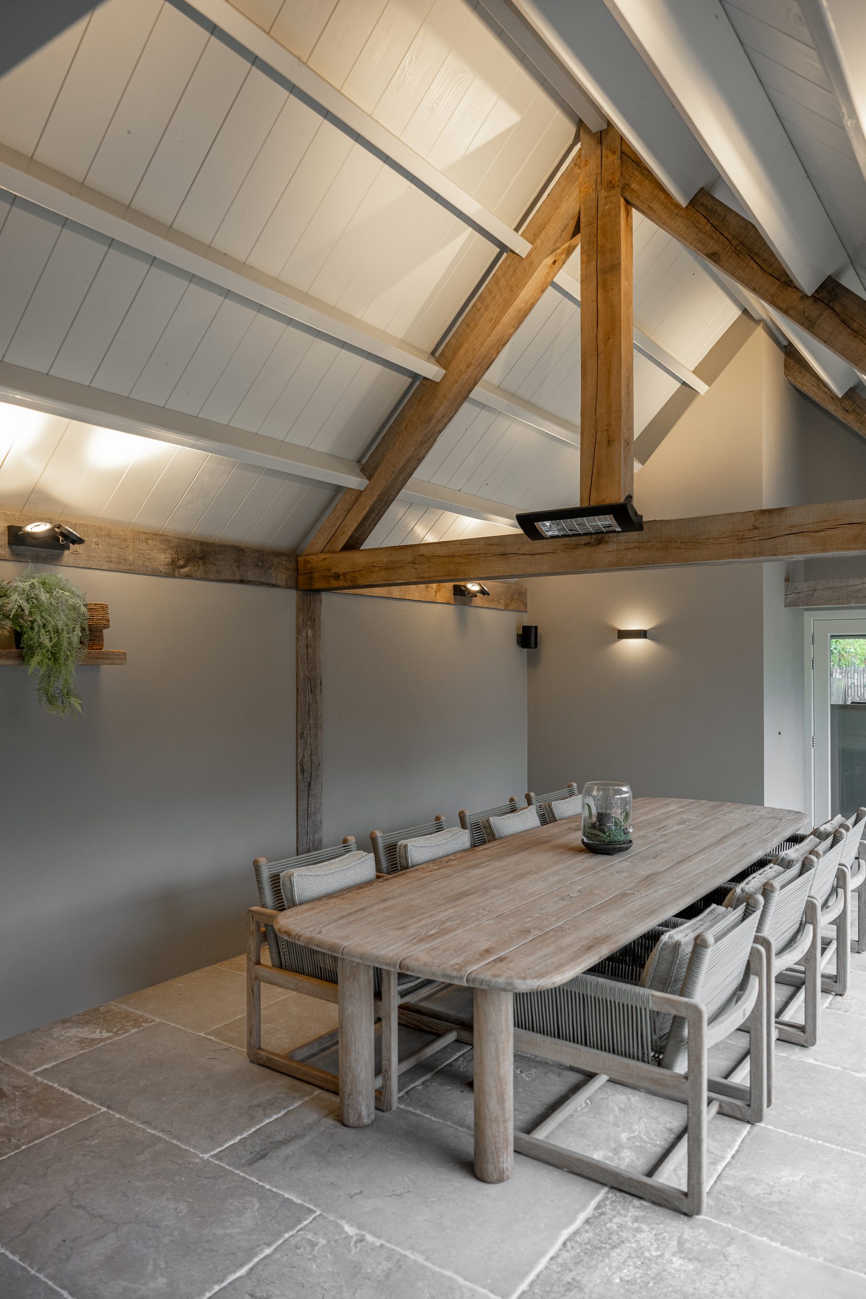 Ruime tuinkamer of overkapping met hoge houten gebinten, een grote houten eettafel en comfortabele tuinstoelen.