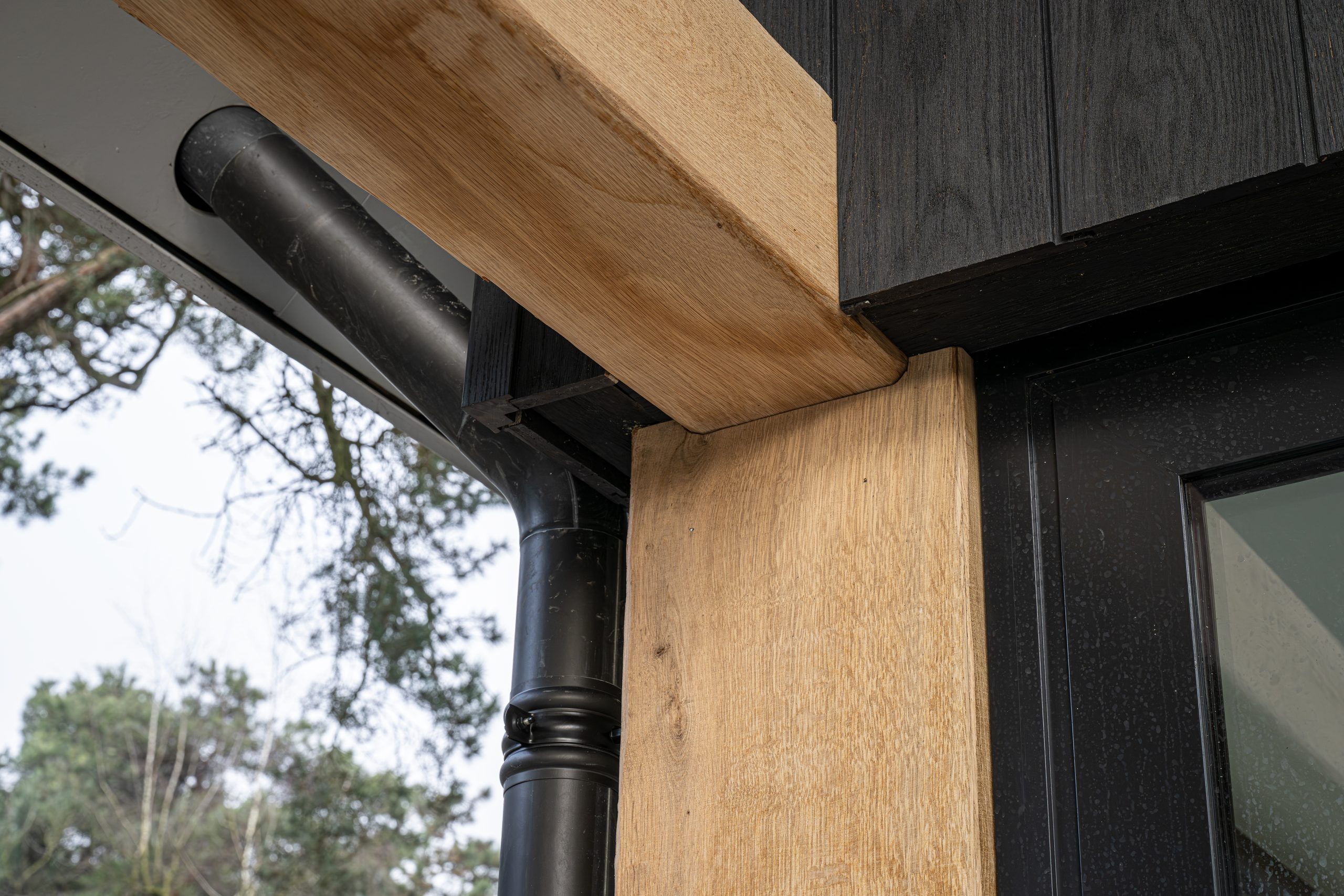 Detail van een strakke houtverbinding van eiken balken bij een zwarte gevel en regenpijp, wat getuigt van vakmanschap.
