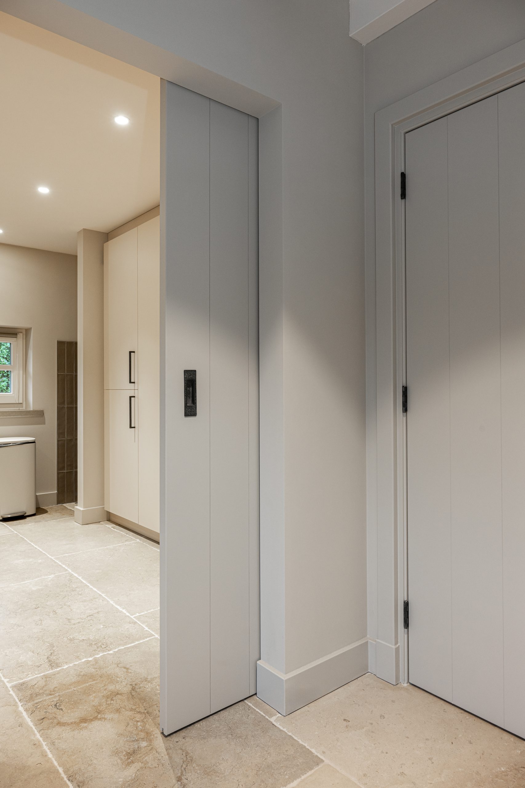 Moderne witte schuifdeur die in de wand verdwijnt (pocket door) met een zwarte inburgeep, grenzend aan een gang met natuurstenen vloer.