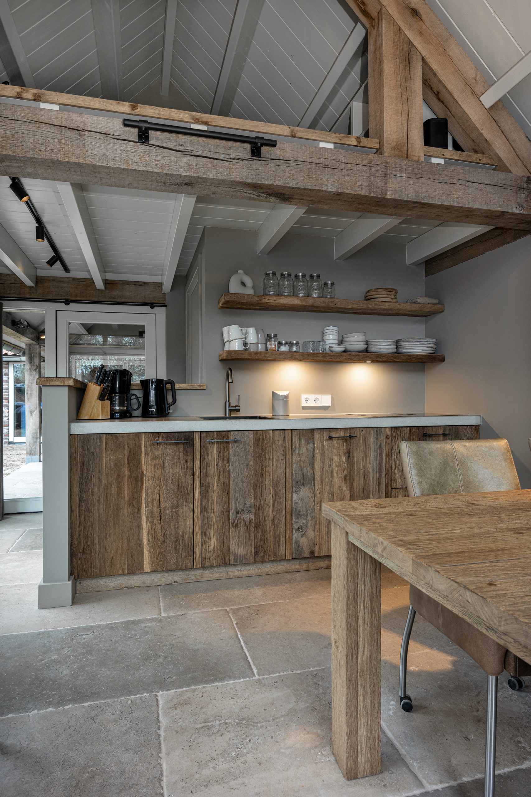 Totaalbeeld van de woonkeuken waarin de robuuste houten dakconstructie en balken zichtbaar zijn boven het keukenblok en de eethoek in tiny house.