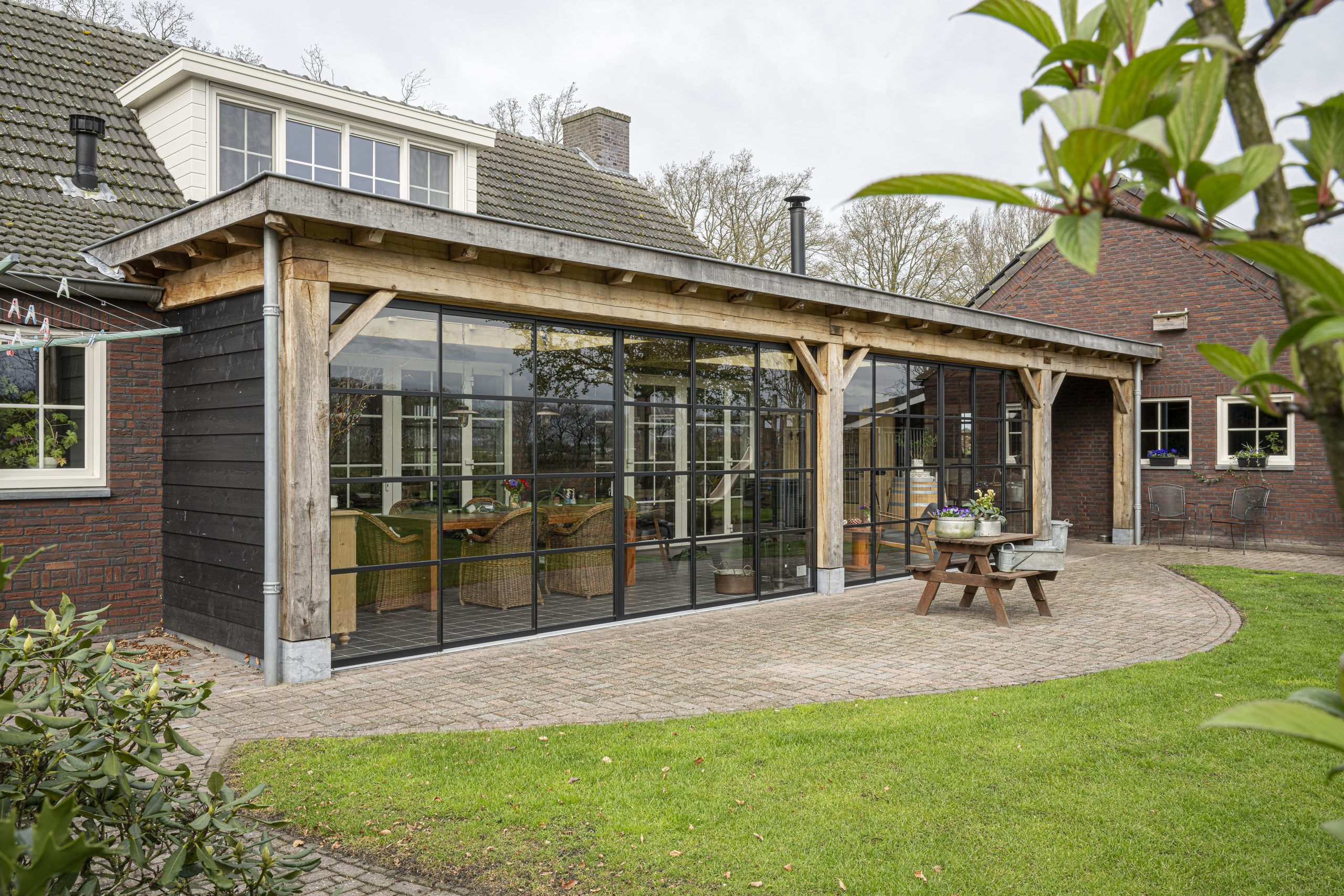 Overzichtsfoto van de achtergevel van de boerderij, uitgebreid met een grote eikenhouten overkapping voorzien van stalen raampartijen.