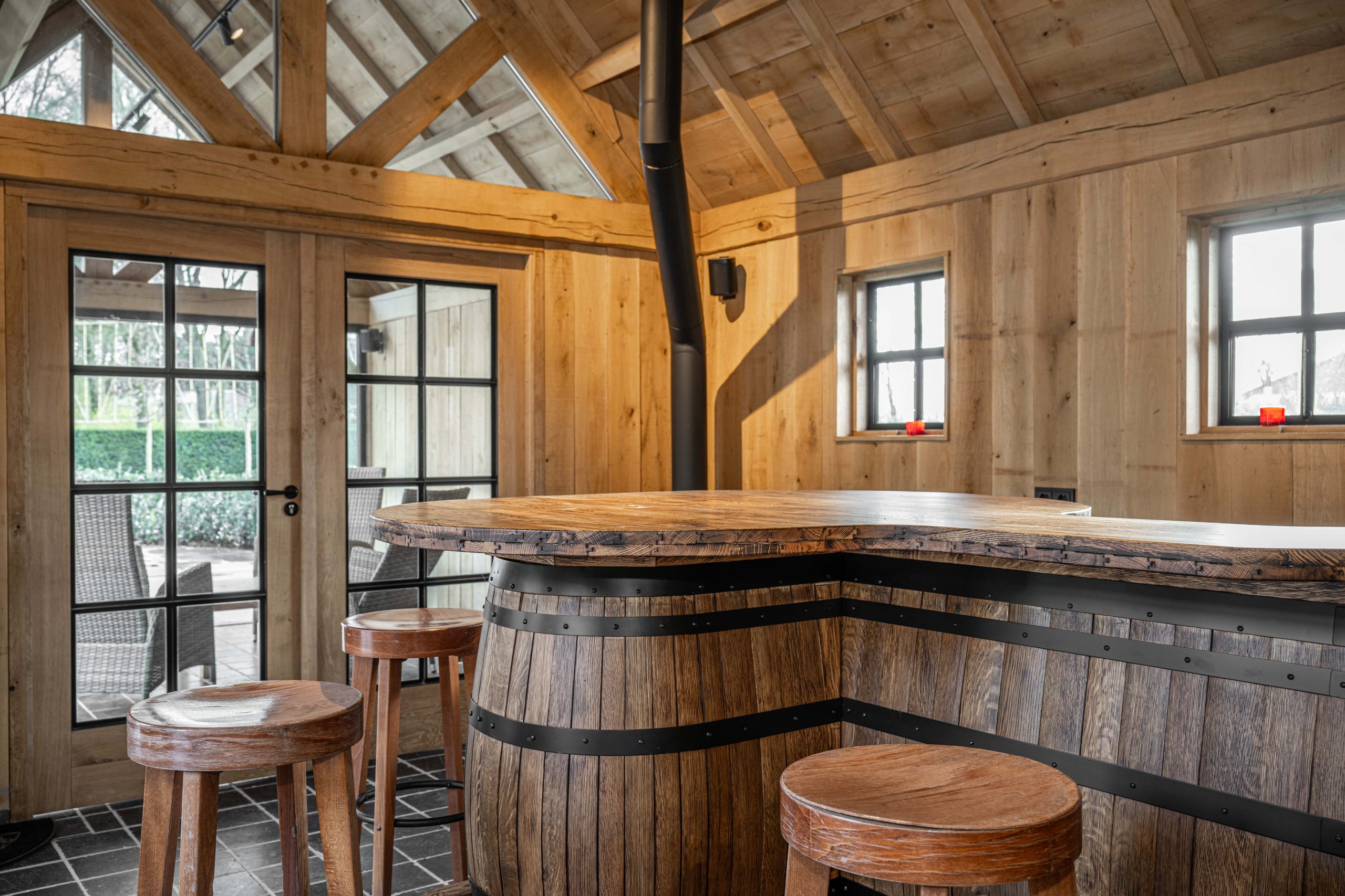 Interieur van een tuinhuis met een bar gemaakt van een houten ton, houten barkrukken en een zwarte houtkachel.