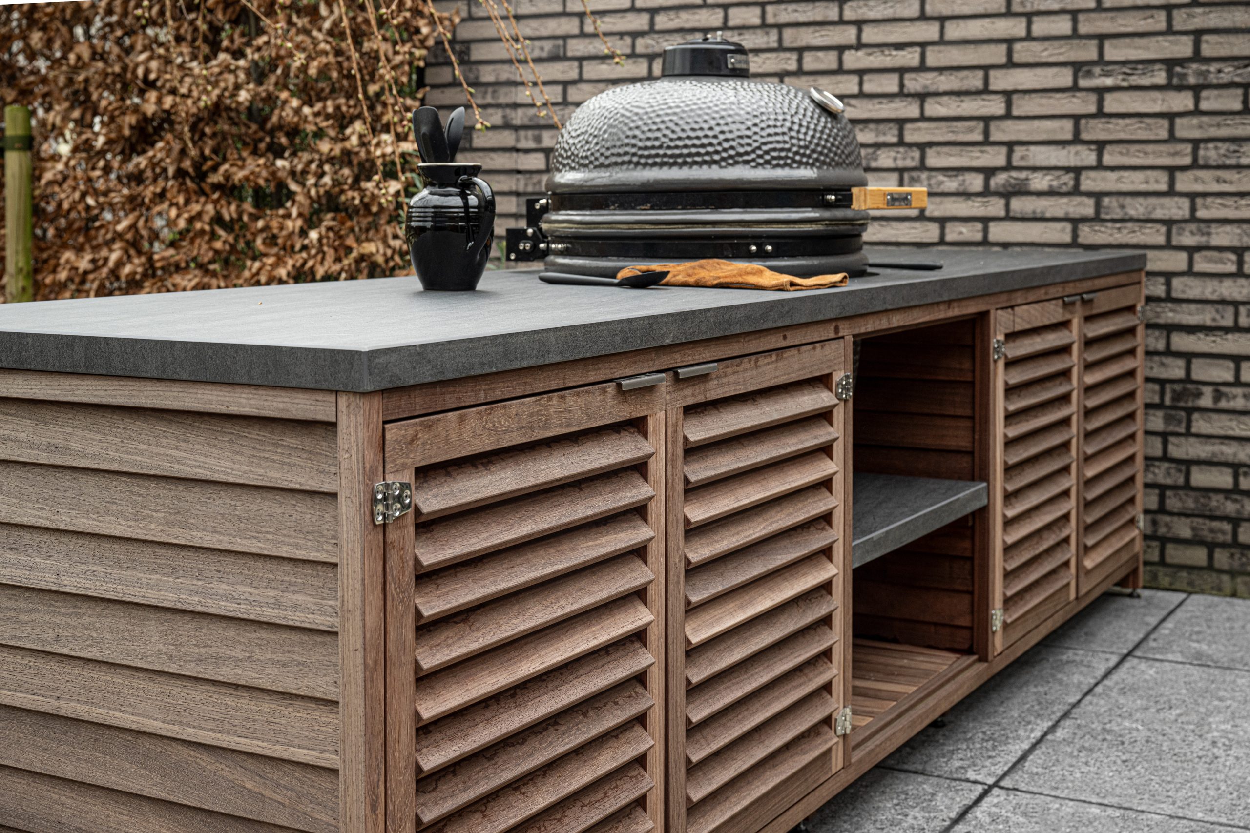 Zijaanzicht van een maatwerk buitenkeuken van padoek hout met een grijs natuurstenen aanrechtblad.