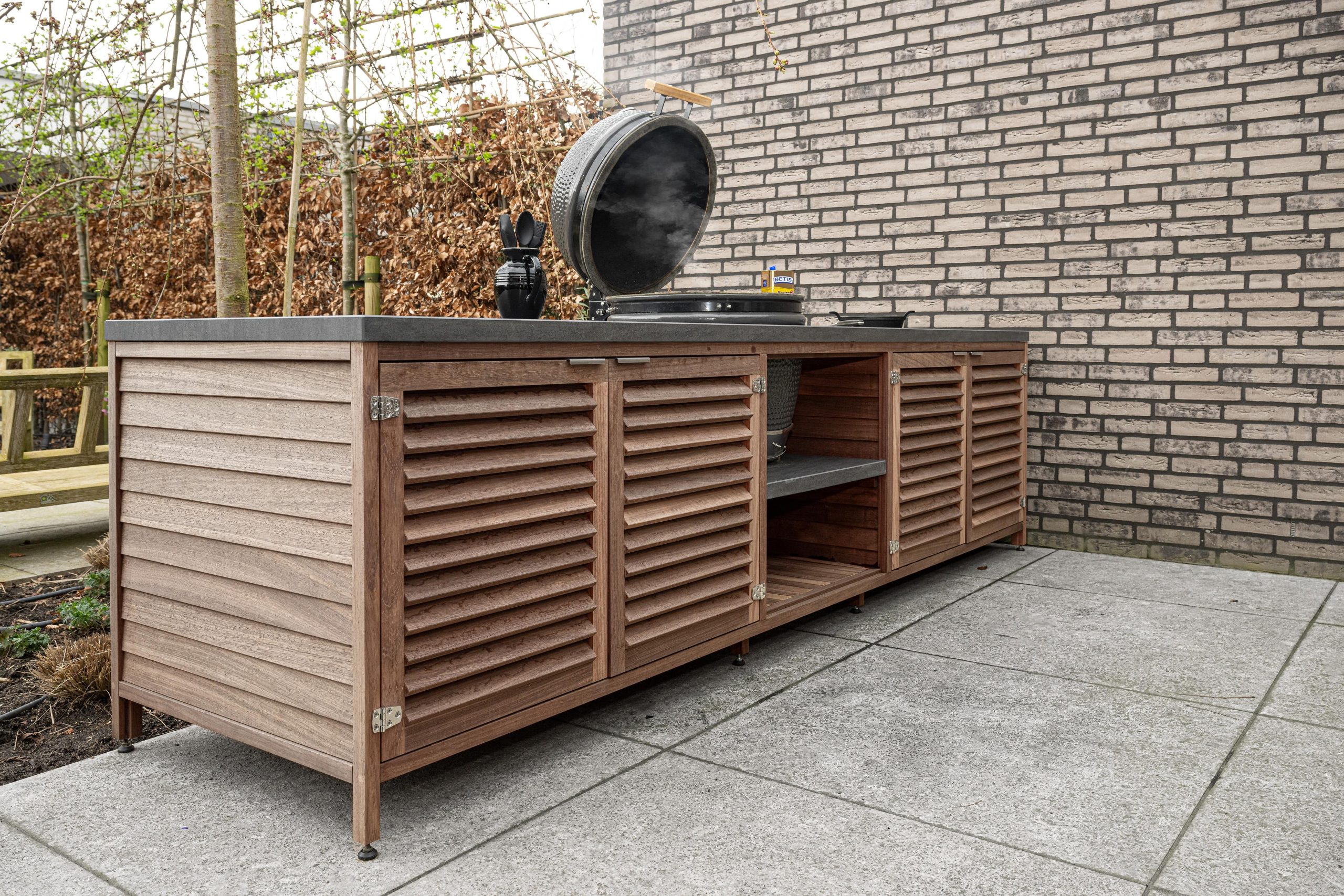 Sfeervol beeld van een padoek houten buitenkeuken in een moderne tuin tegen een bakstenen muur.