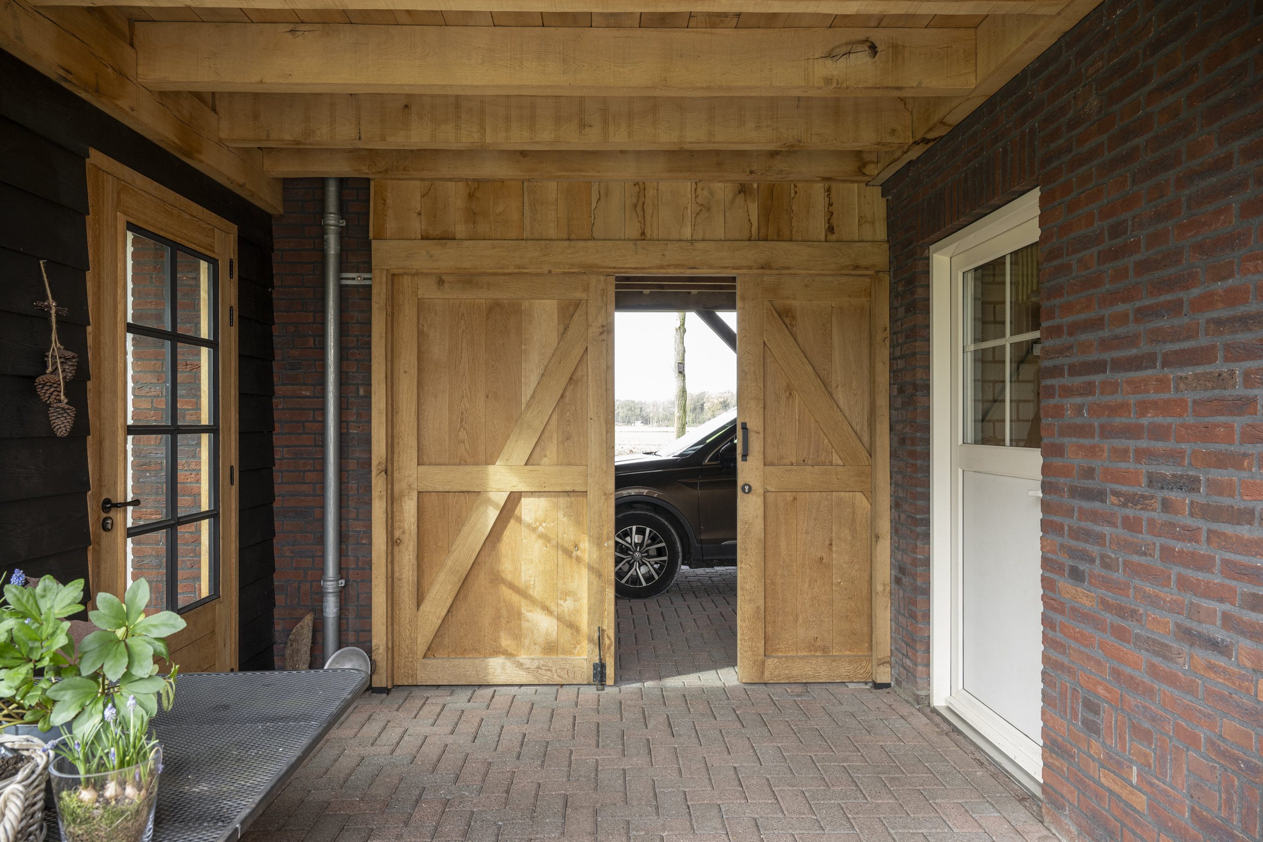Zicht onder de carport waar de grote eikenhouten schuifdeuren op een kier staan en een zwarte auto zichtbaar is in de garage erachter.