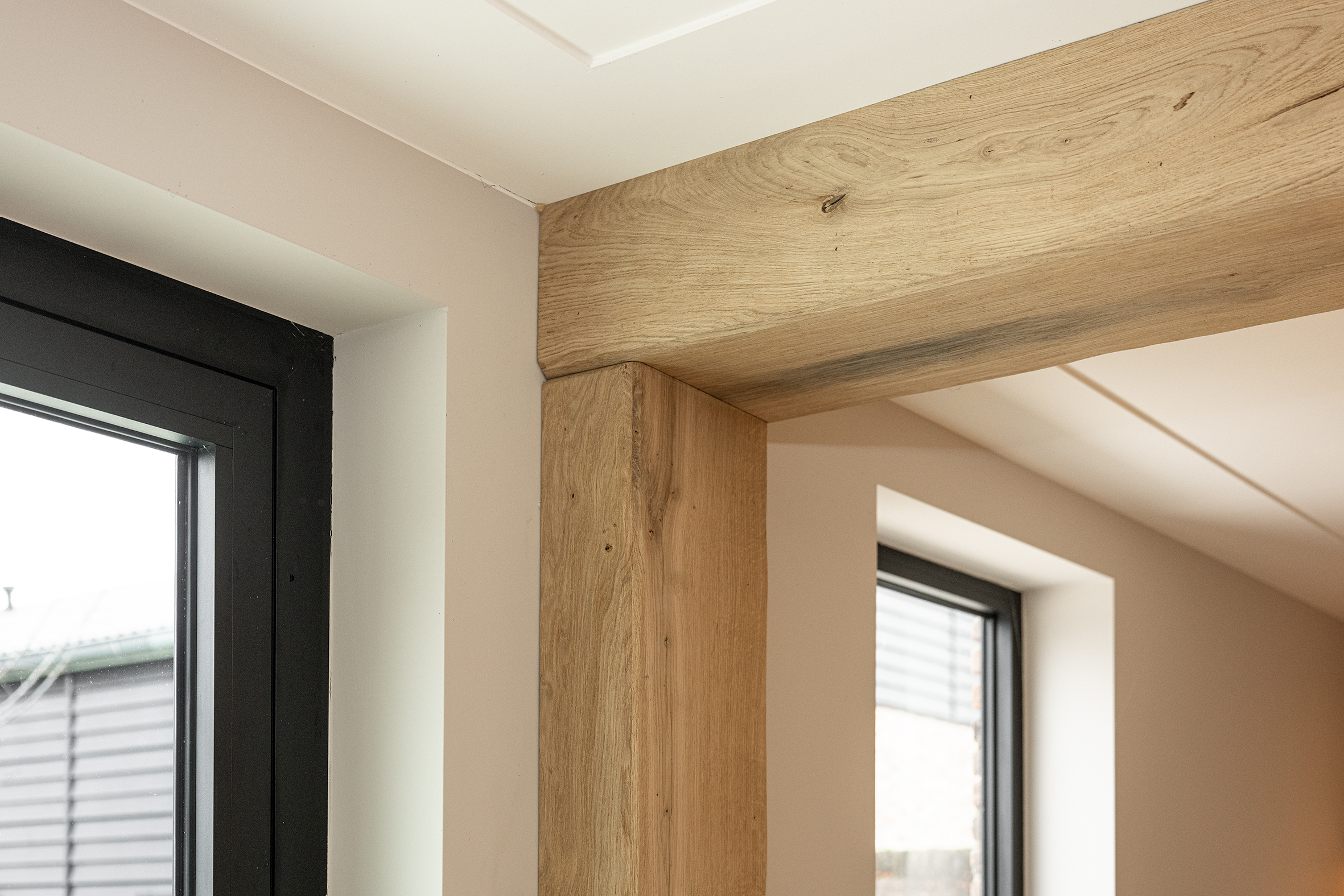 Detailopname van de haakse verbinding tussen een verticale en horizontale eikenhouten balk naast een zwart raamkozijn.