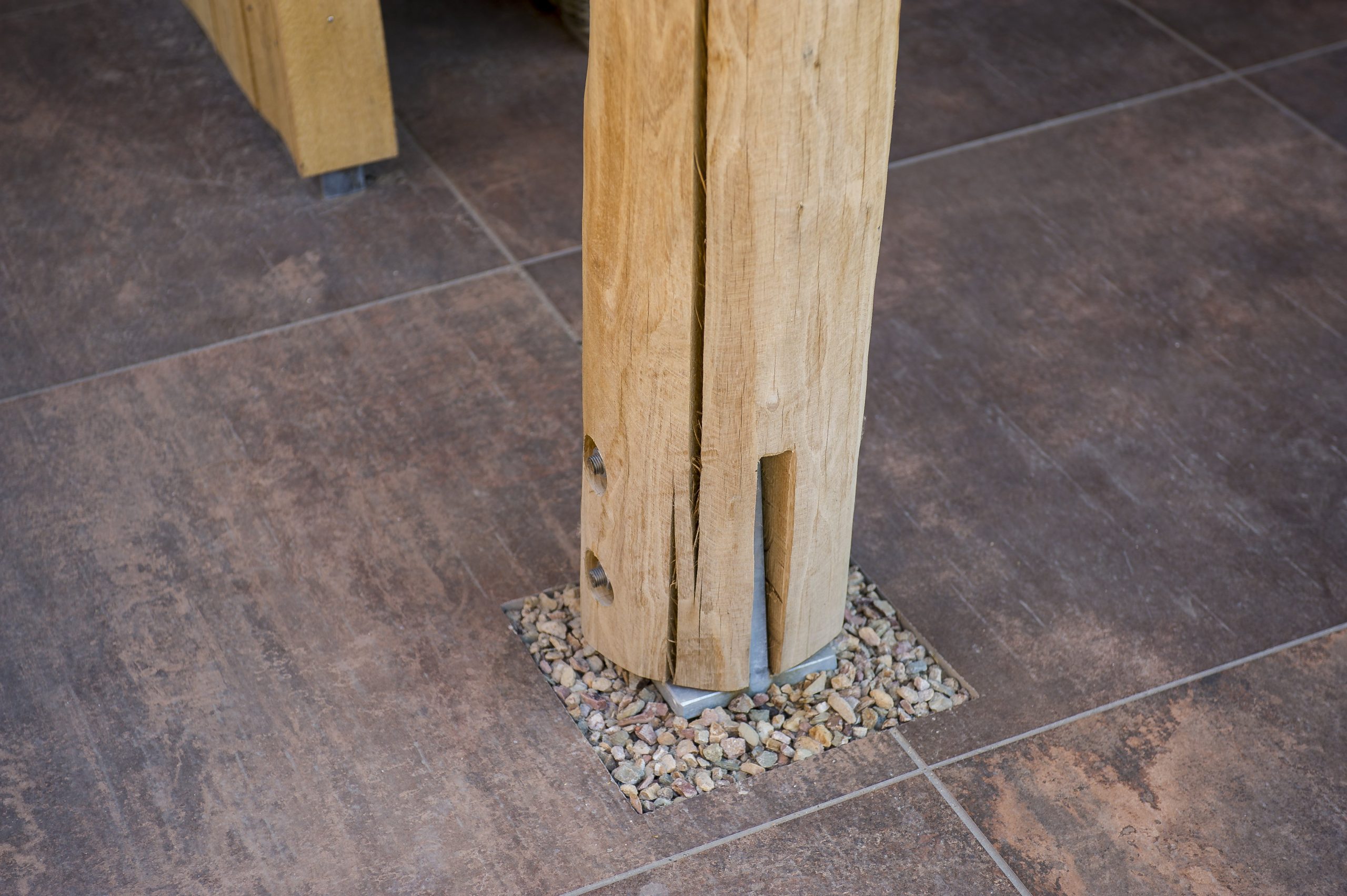 Close-up van de onderzijde van een robuuste eikenhouten staander van een overkapping, gemonteerd op een metalen voet omringd door grind in een tegelvloer.