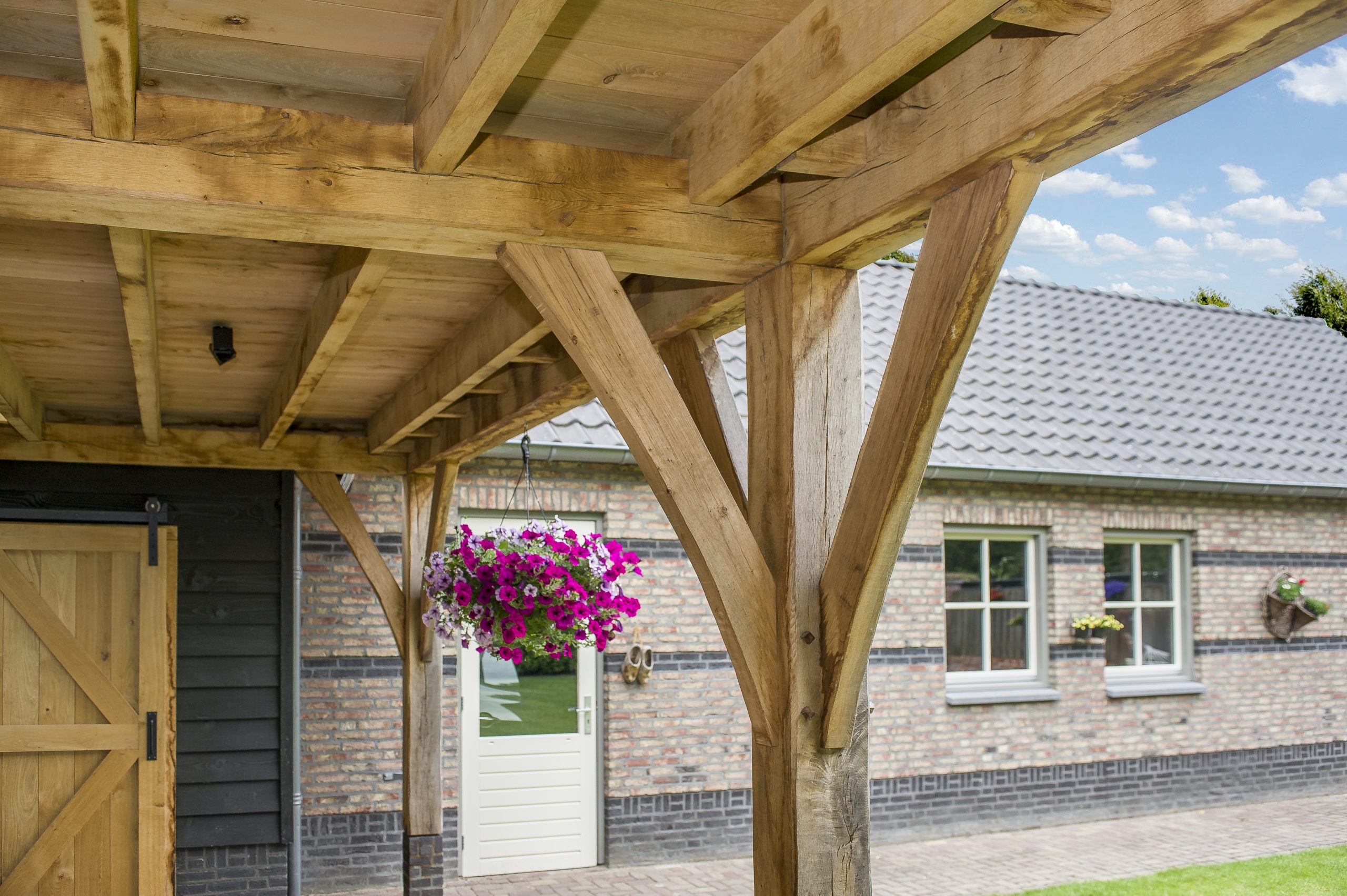 Close-up van de ambachtelijke houtverbindingen en schoren van een eikenhouten overkapping, met op de achtergrond een hangende bloempot met paarse petunia's.