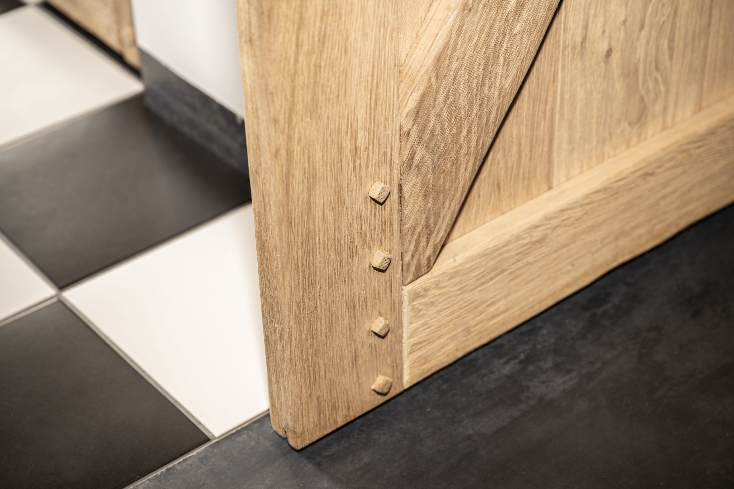 Close-up van de hoek van een massief houten deur met traditionele houten pennen (doken) op een zwart-witte tegelvloer.