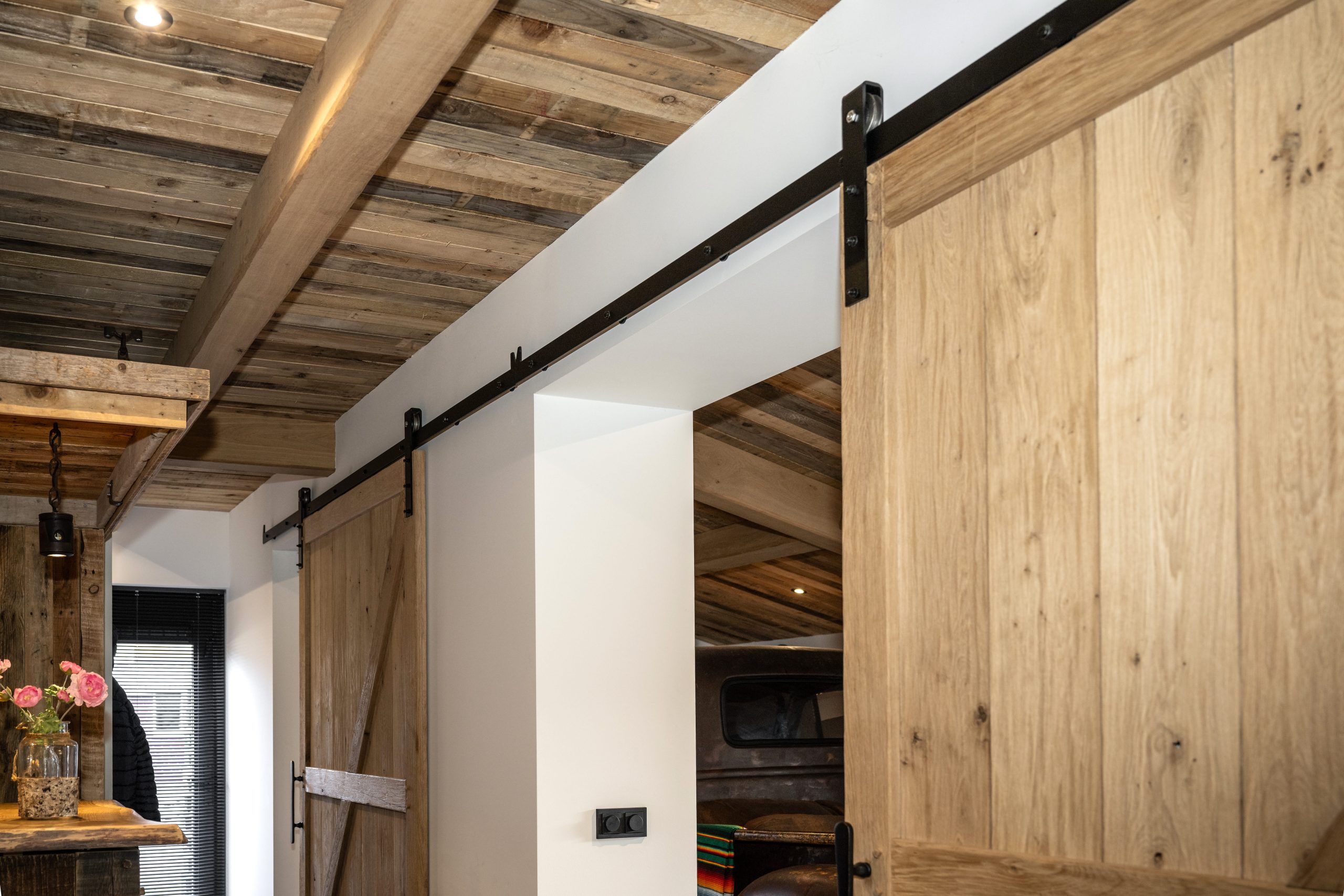 Detail van een zwart metalen railsysteem voor een robuuste houten staldeur tegen een wit plafond met eiken balken.