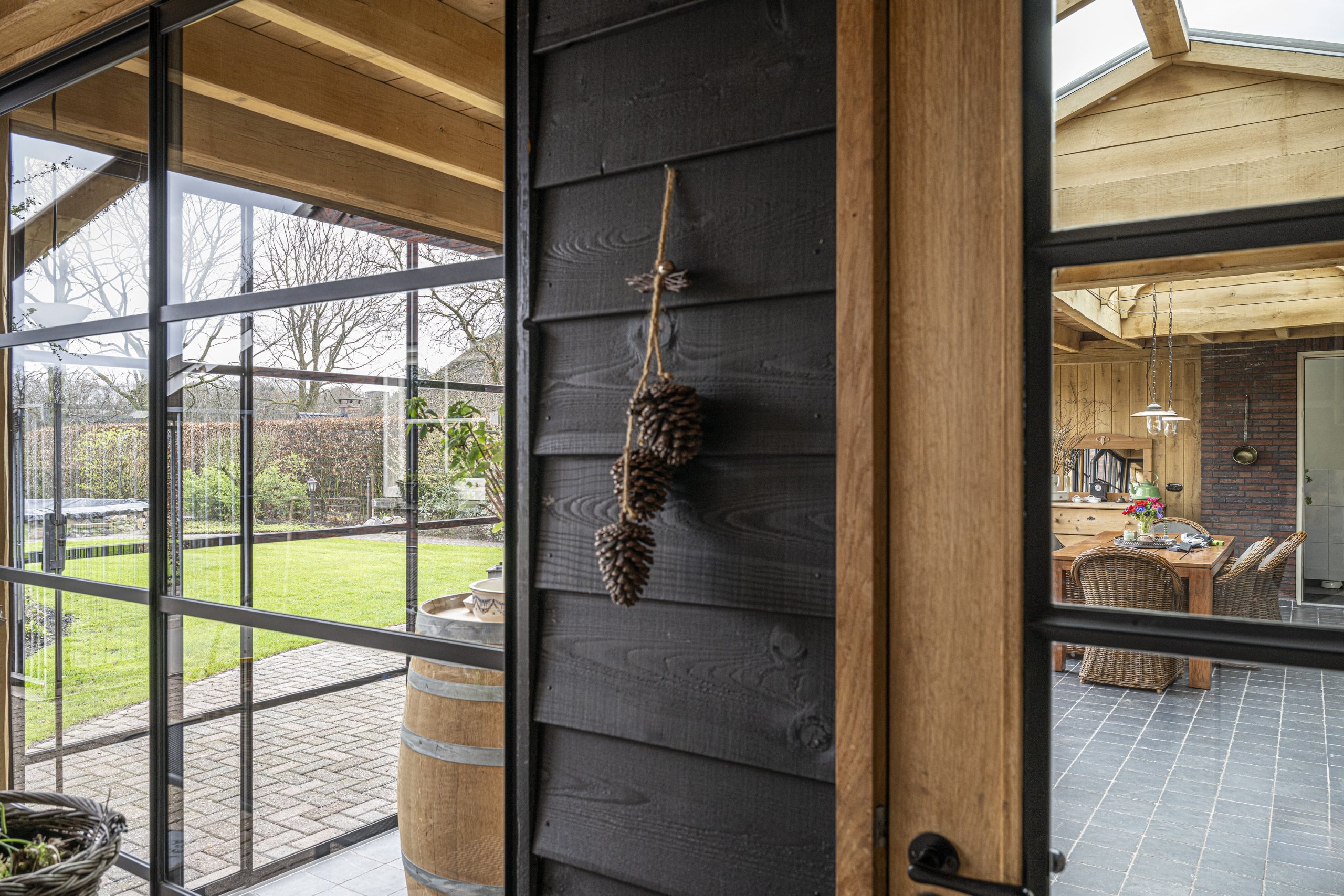 Detailopname van binnenuit kijkend door een geopende eikenhouten binnendeur en een gesloten zwarte stalen deur met glas naar het terras.