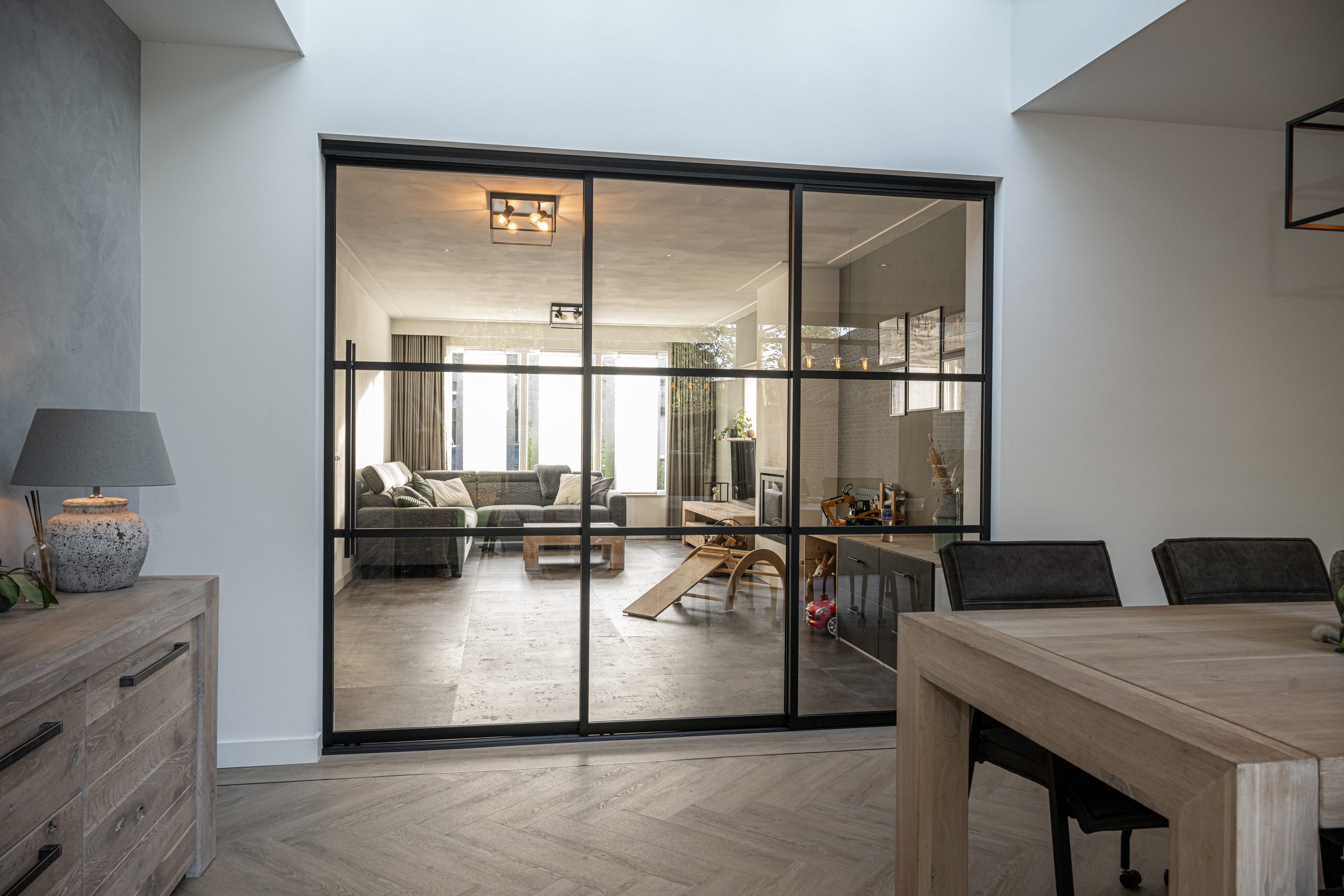Zicht vanuit een lichte eetkamer door een glazen stalen pui naar een woonkamer met grijze bank.