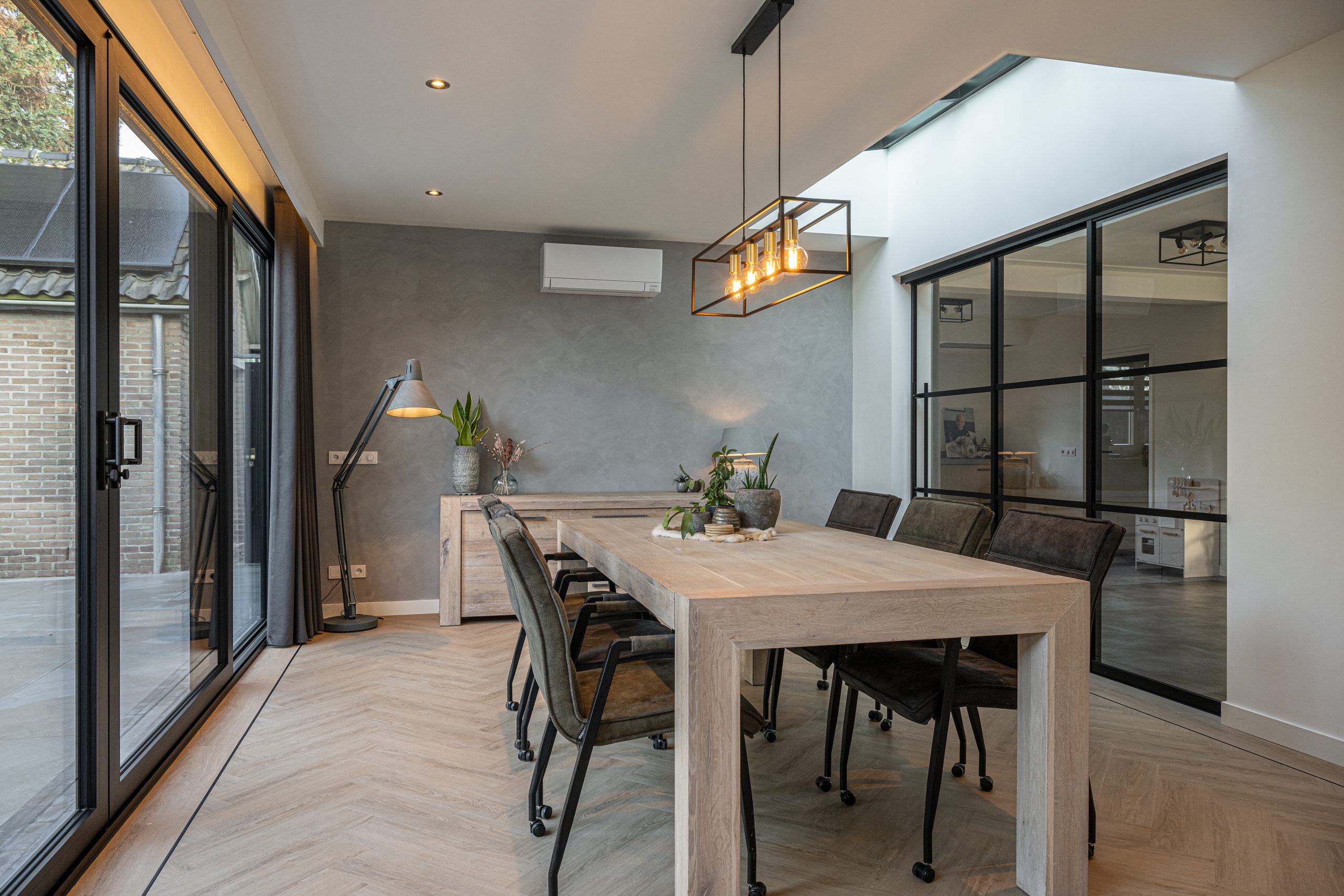 Interieur van een eetkamer met een grijze betonlook wand, een robuuste houten tafel en een moderne hanglamp.