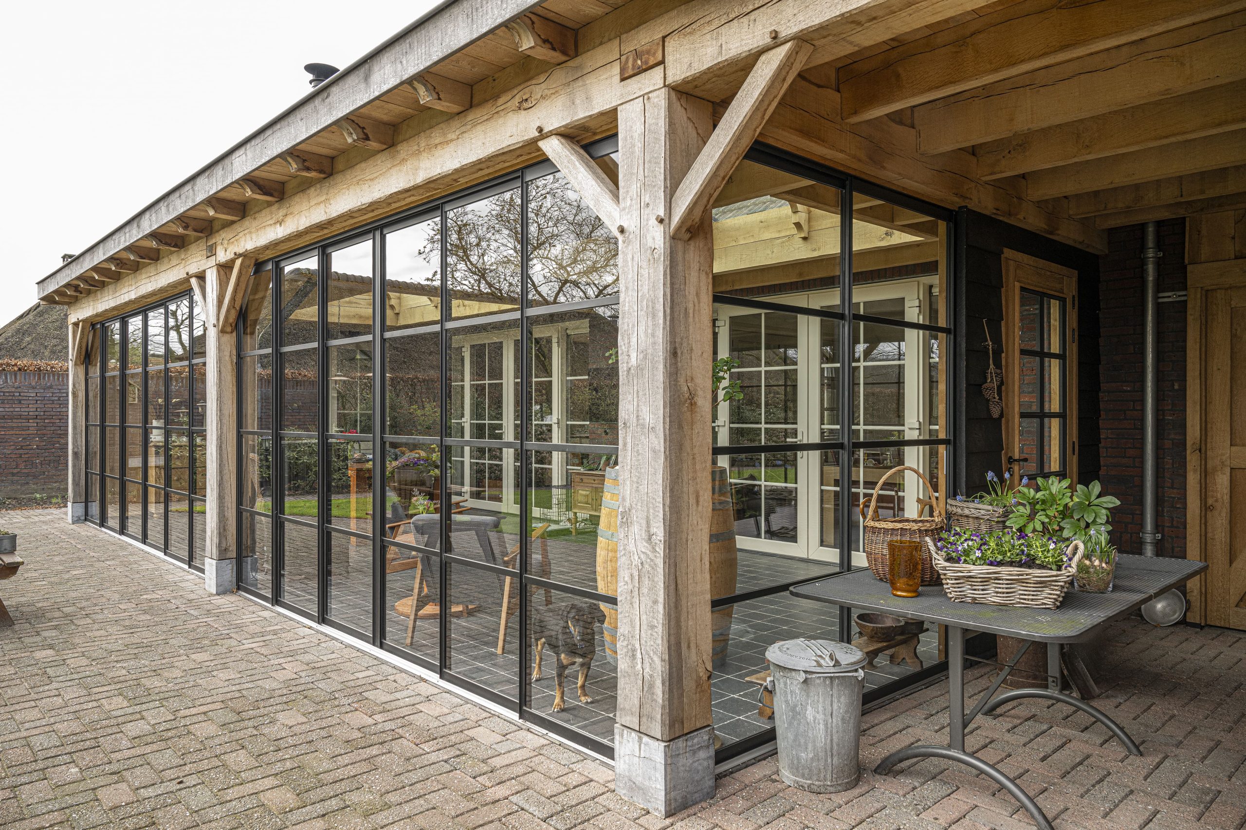 Buitenaanzicht van de hoek van de eikenhouten overkapping, gekenmerkt door zwarte houten gevelbekleding en grote stalen ramen met roeden.