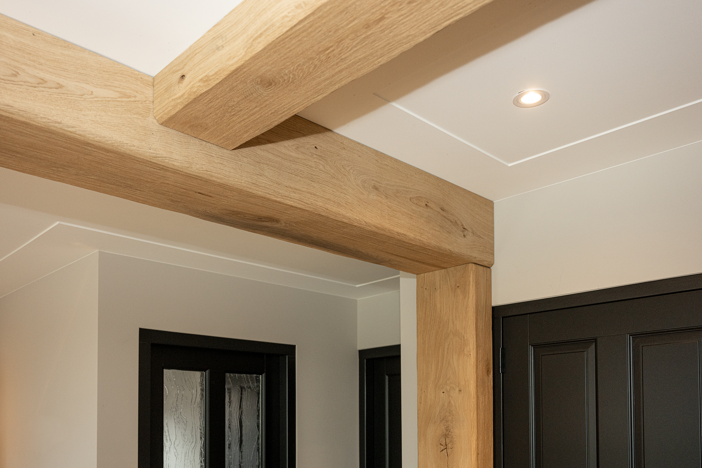 Plafonddetail van robuuste eikenhouten balken gecombineerd met een strak wit stucwerk plafond en inbouwspots.