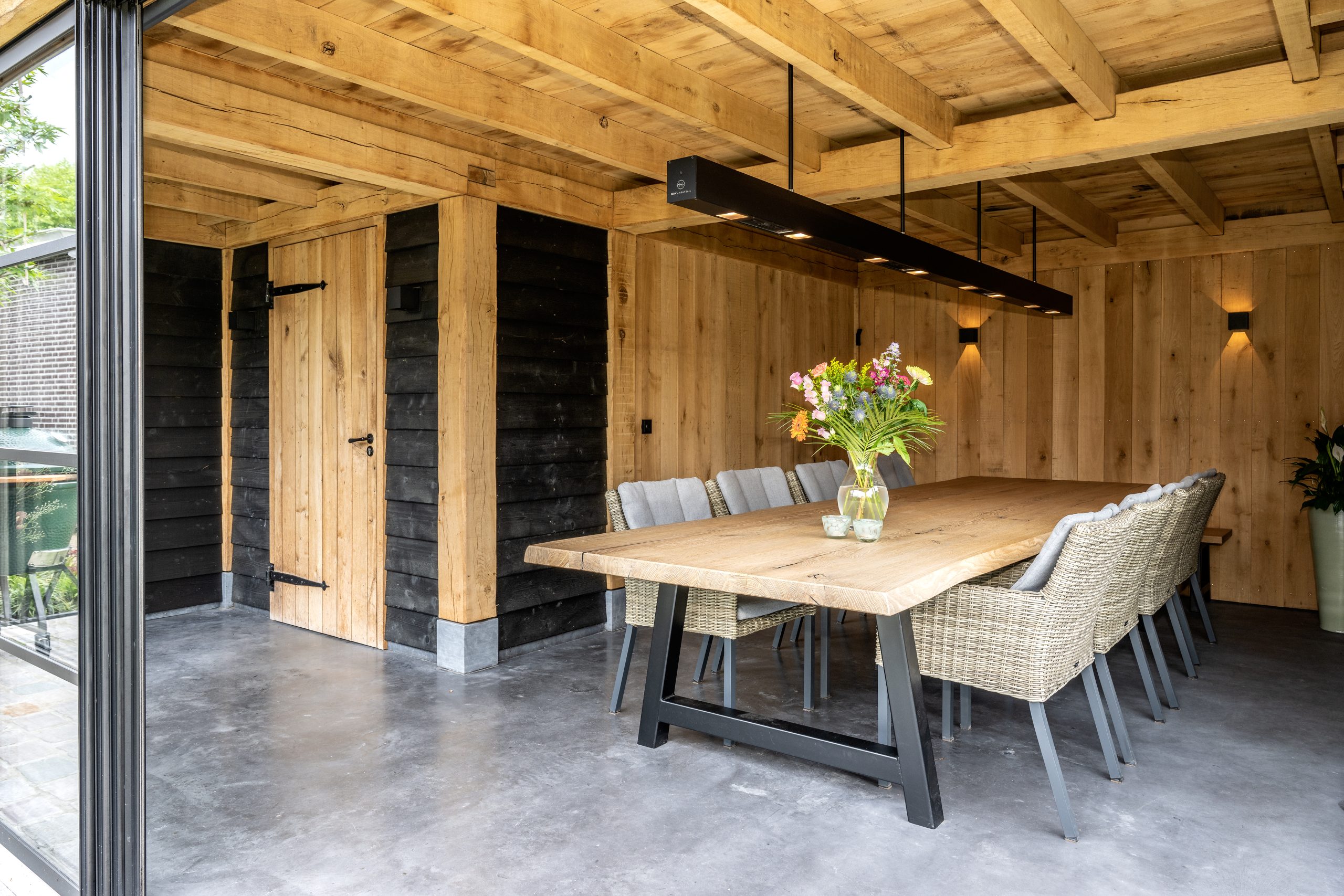 Een lange houten eettafel met gevlochten stoelen en moderne hangverlichting in een tuinhuis.