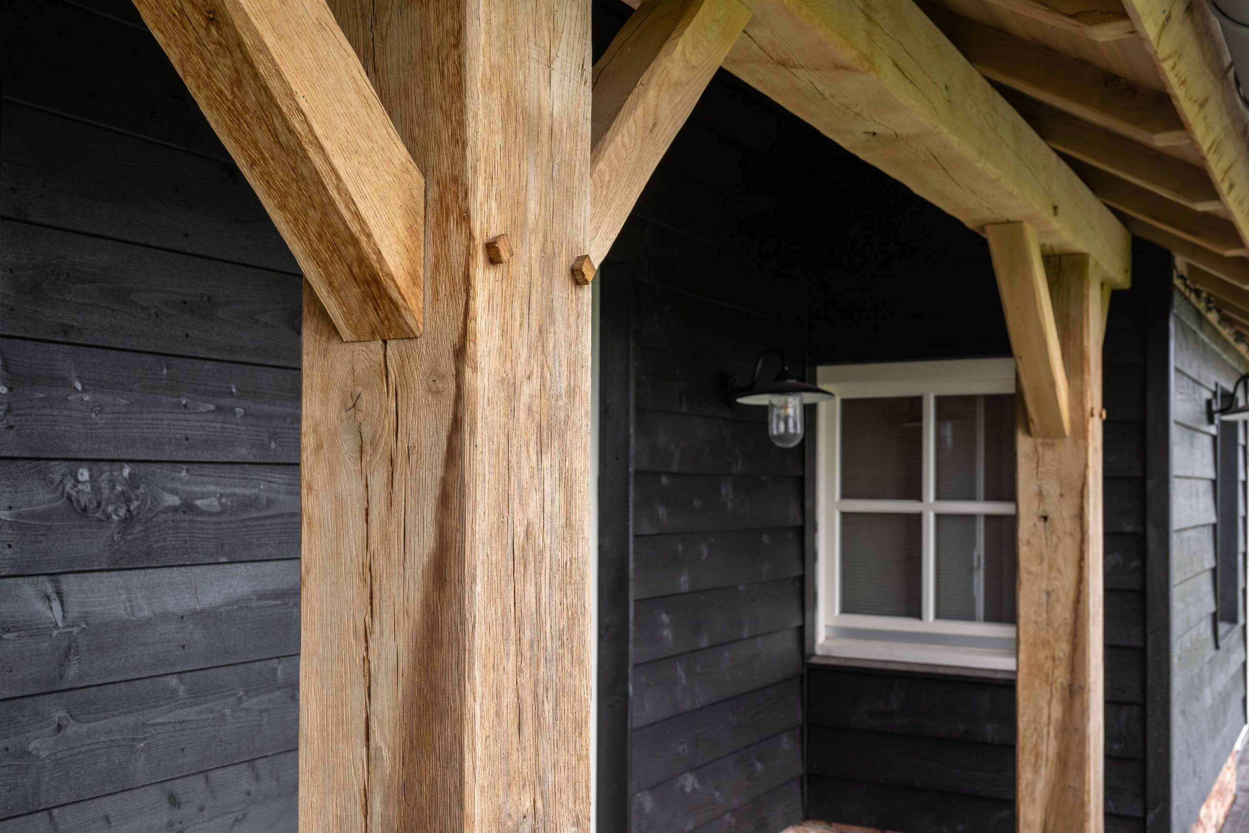 Close-up van de constructie van een eikenhouten overkapping met traditionele schoren tegen een zwarte houten wand.