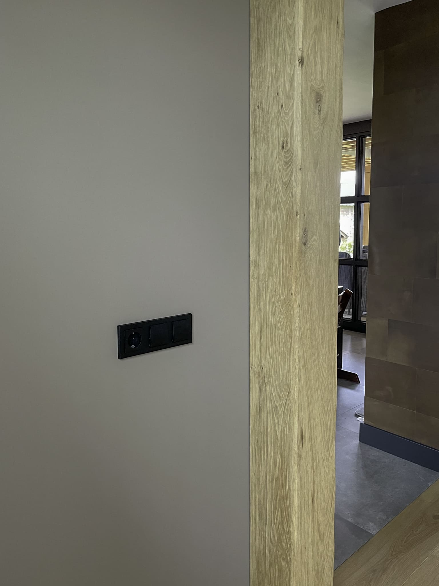 Detailopname van een massief eikenhouten balk naast een strakke wand met zwart modern schakelateriaal.