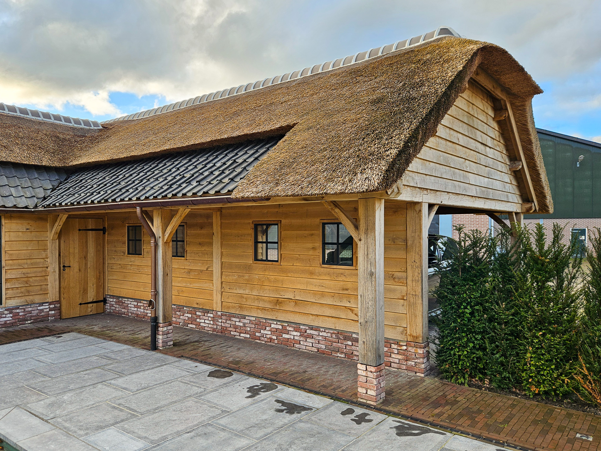Zijaanzicht van een langgerekt eikenhouten bijgebouw met een rieten kap en zwarte raamkozijnen.
