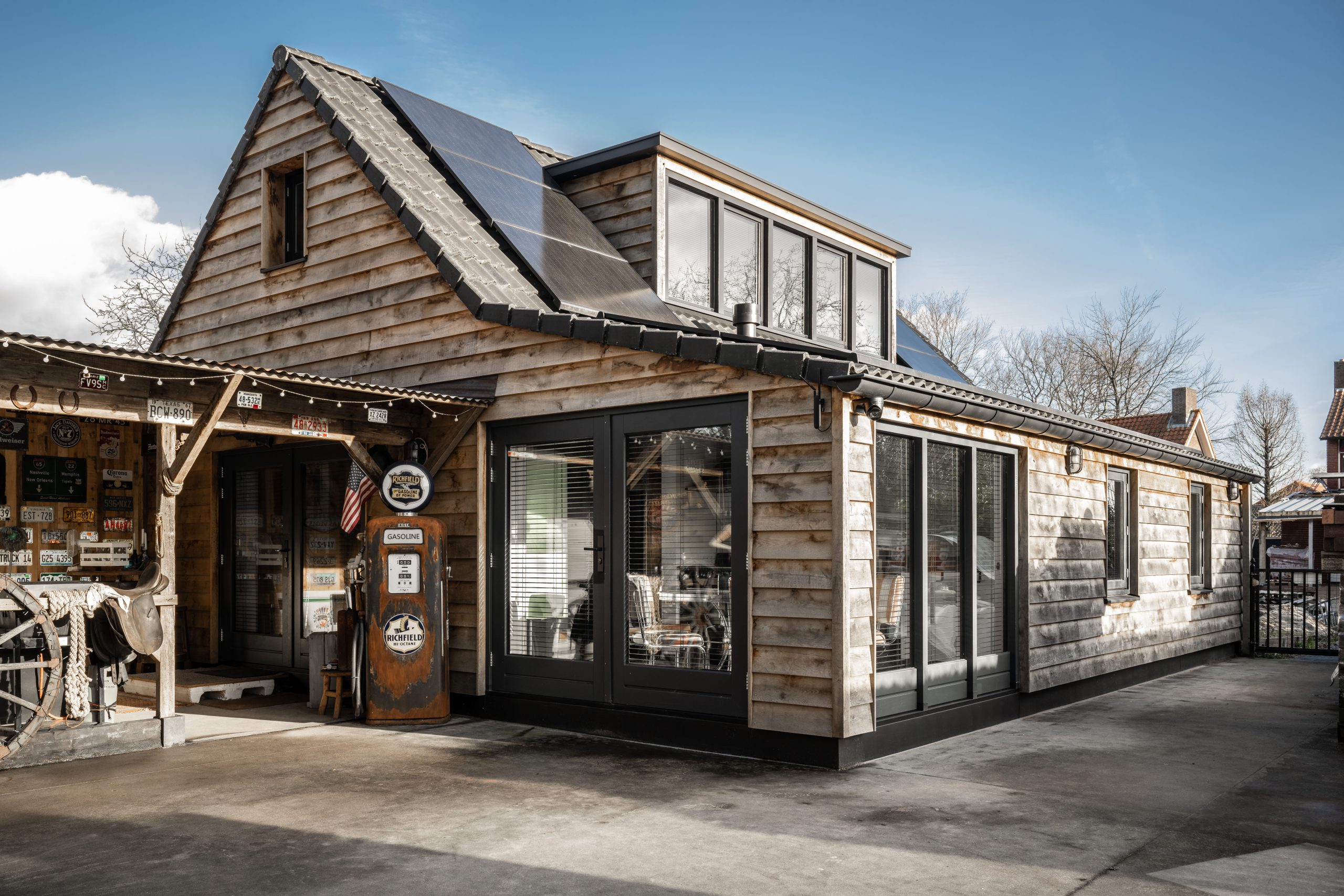 Exterieur van een houten gebouw met zonnepanelen, een vintage Richfield benzinepomp en een veranda met Amerikaanse nummerplaten.