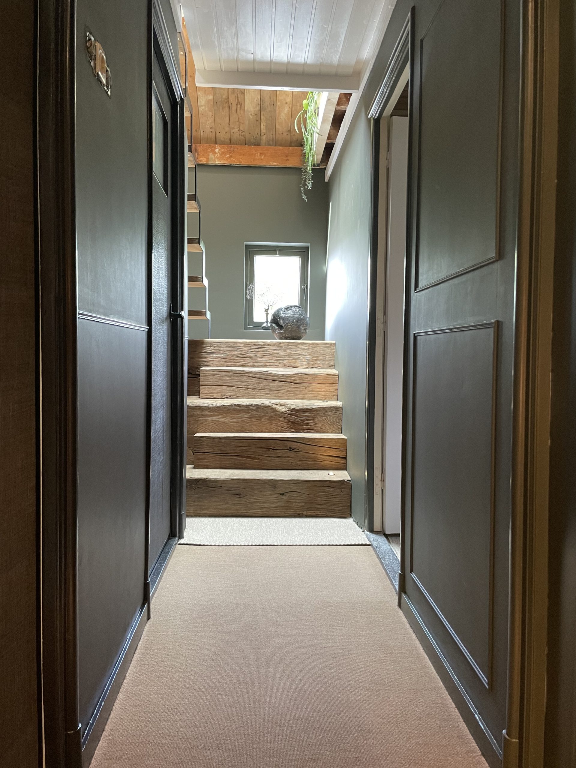 Doorkijkje door een donkere gang naar een robuuste trap van massieve eiken blokken met een klein raam op de achtergrond.