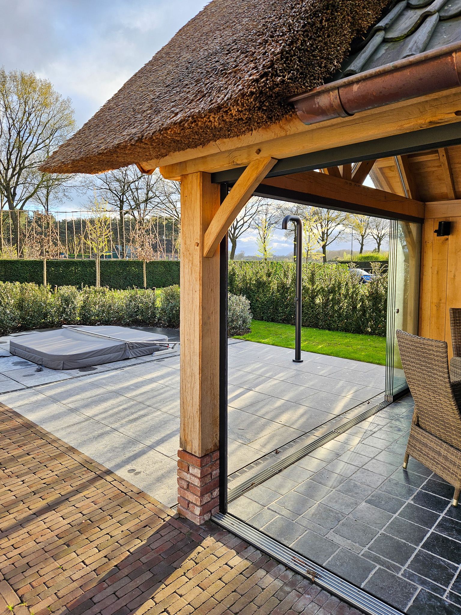 Zicht vanuit een tuinhuis door een geopende glazen schuifwand naar een terras met een afgedekte jacuzzi.