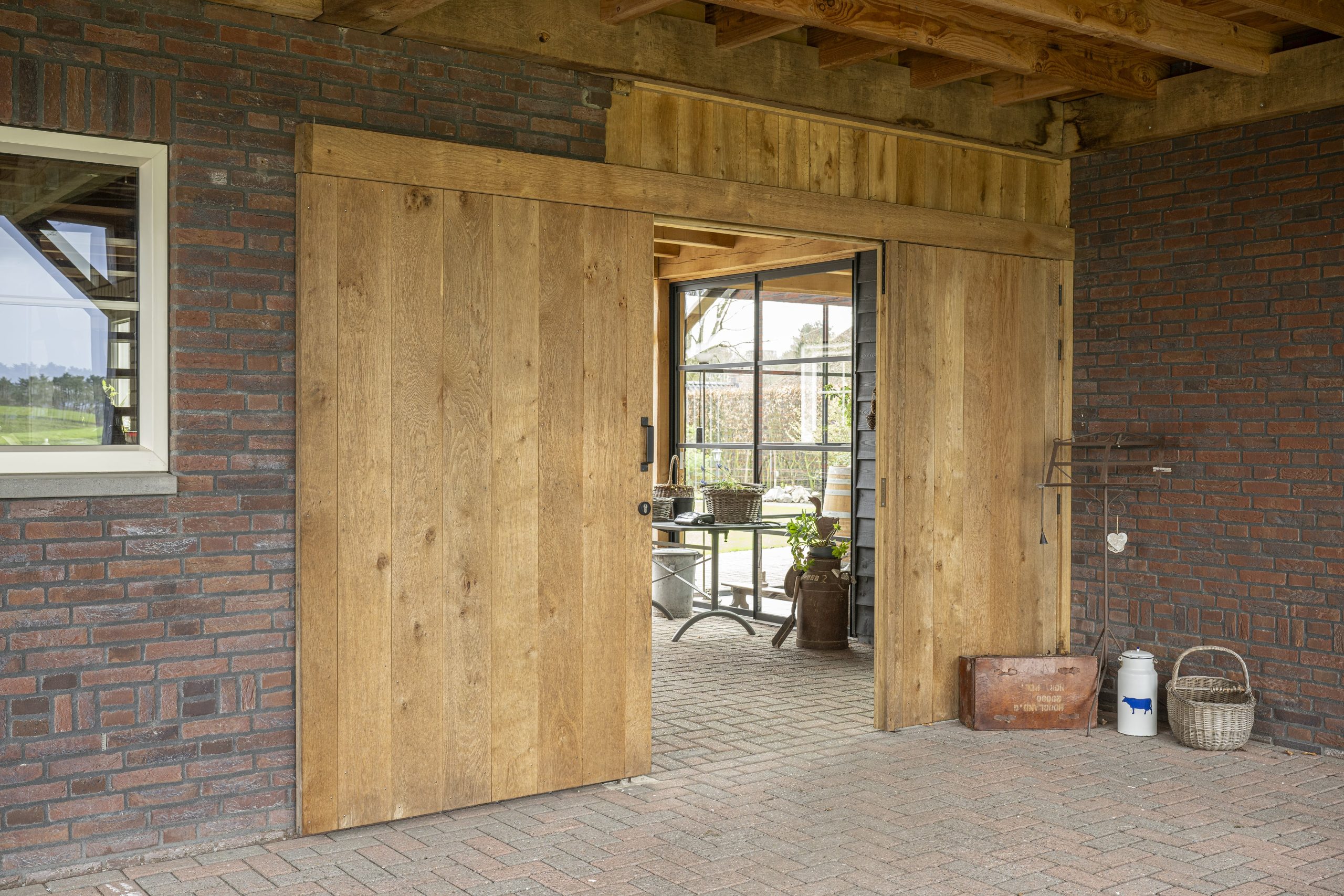 Een brede dubbele schuifdeur van eikenhout, volledig gesloten, geïntegreerd in de bakstenen buitenmuur van het bijgebouw.