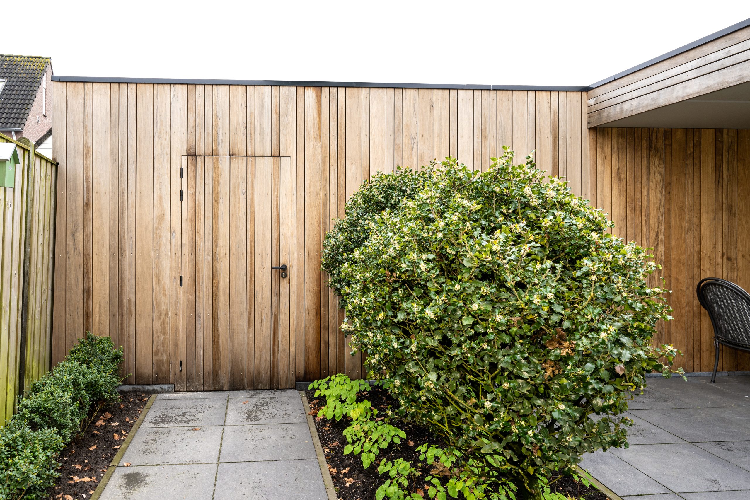 Detail van een houten wand met een naadloos weggewerkte deur en een groene struik op de voorgrond.