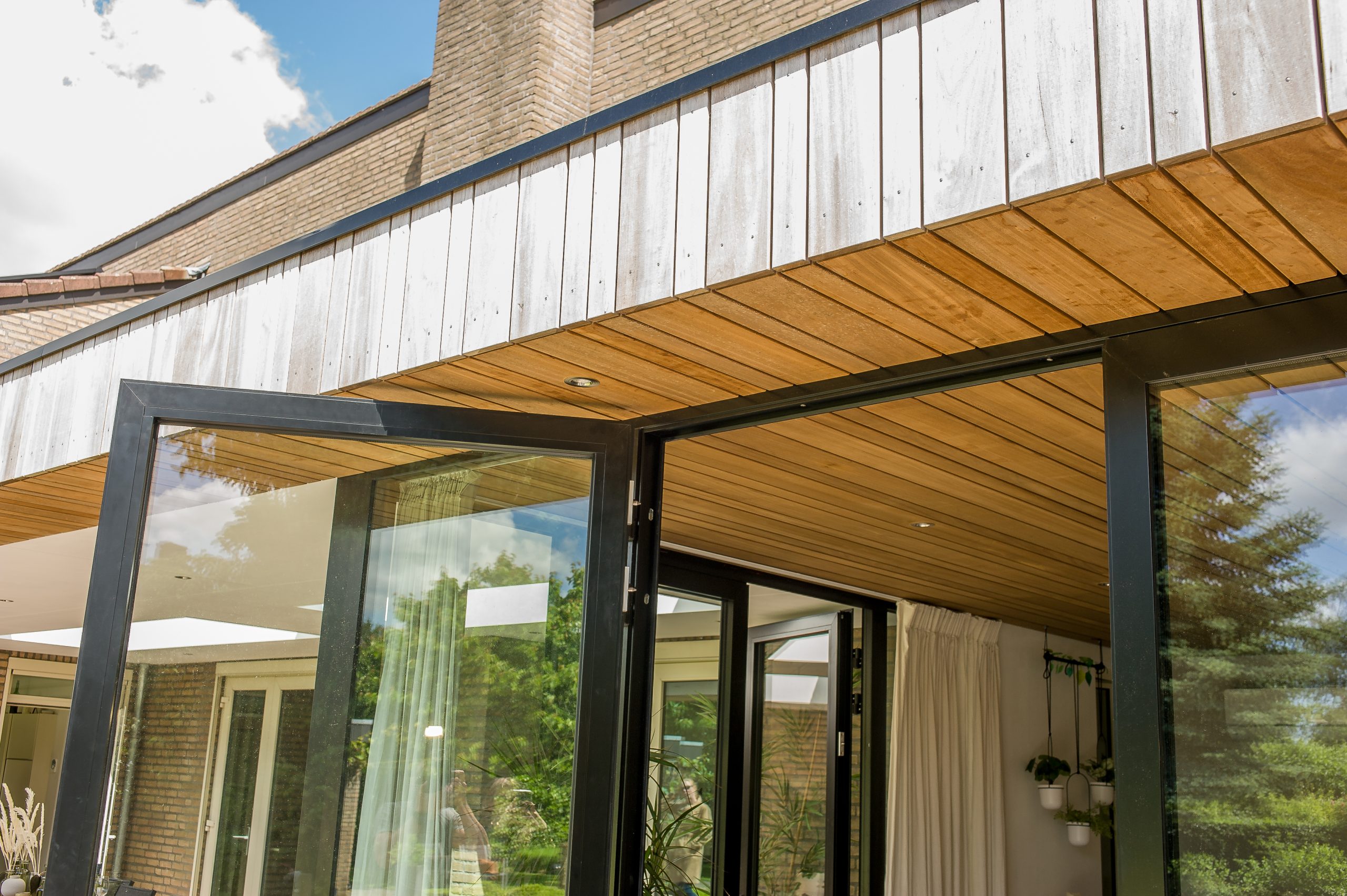 Close-up van de houten gevelbekleding en het boeiboord van een overkapping met zwarte aluminium openslaande deuren.