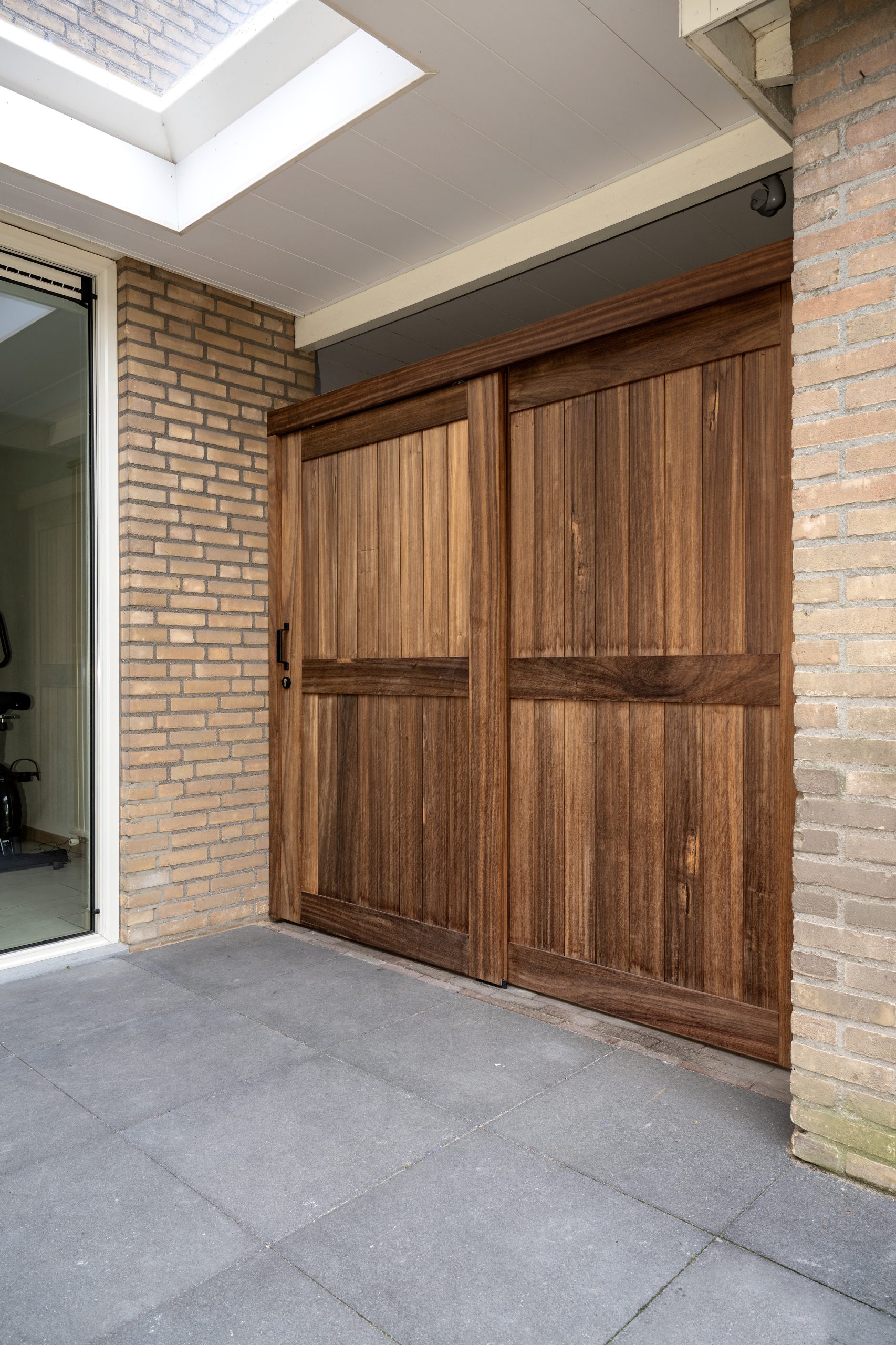 Een robuuste houten schuifdeur gemonteerd tegen een lichtbruine bakstenen muur op een betegeld terras.