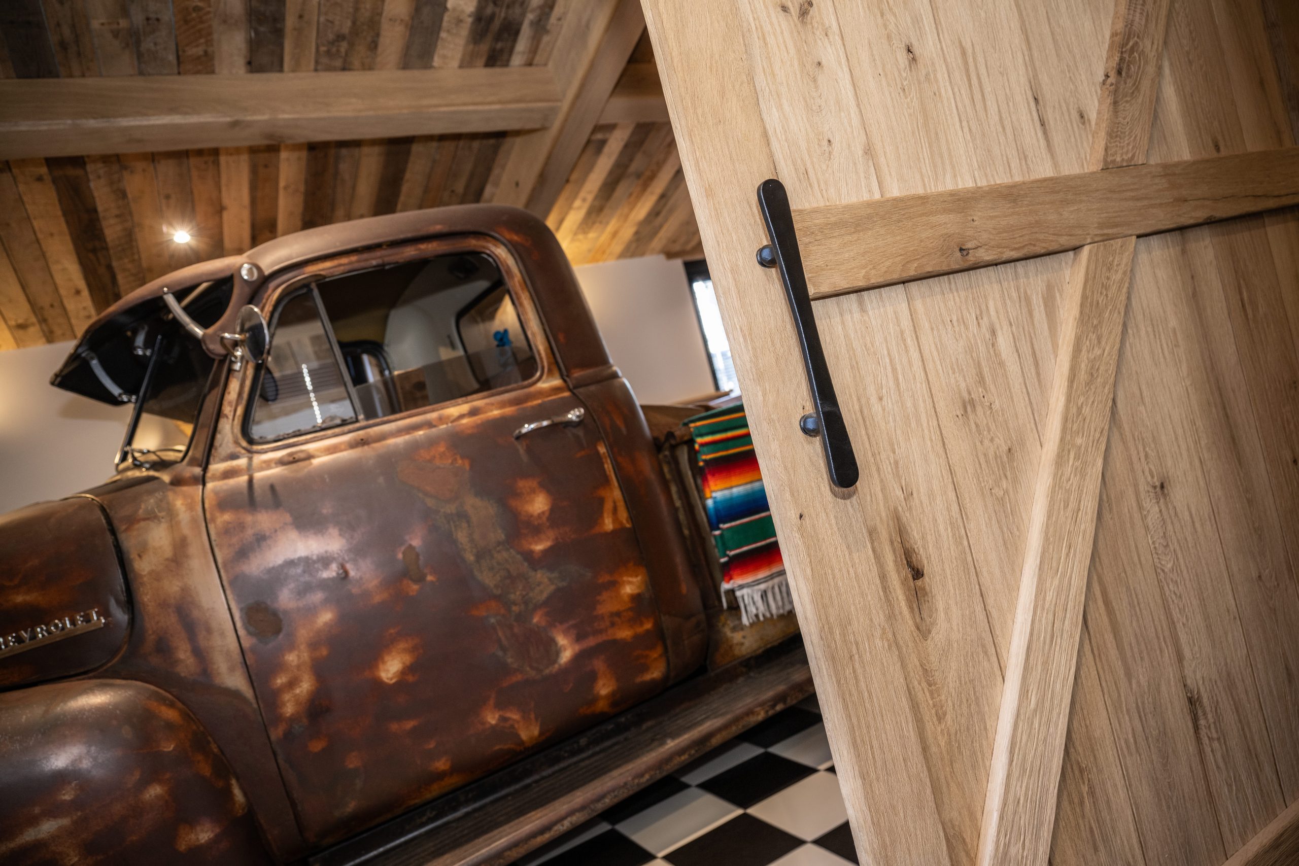 Een robuuste houten staldeur met een lange zwarte handgreep die zicht biedt op een vintage, roestige Chevrolet pick-up truck.