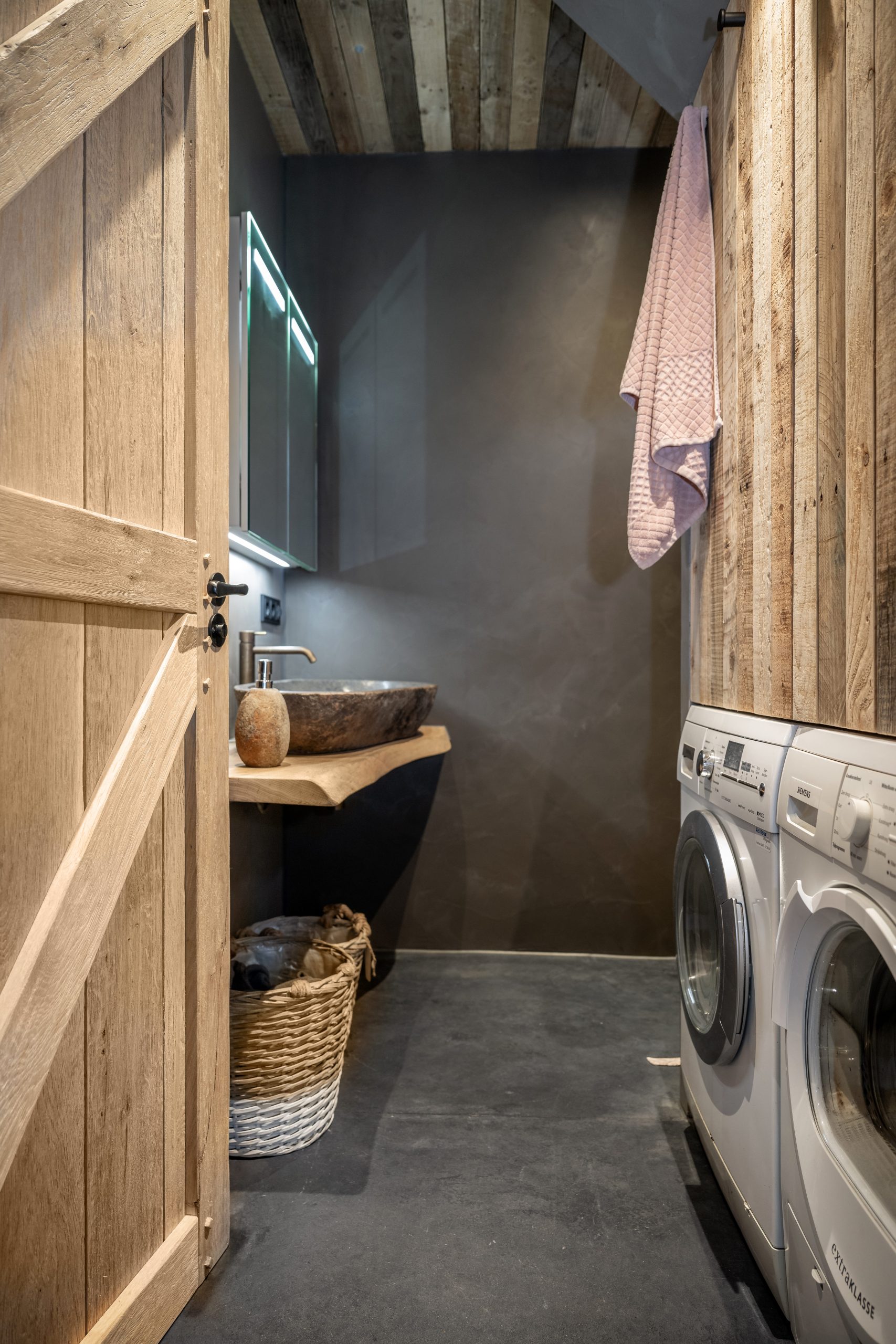 Badkamer met een natuurstenen wasbak op een boomstam-blad, betonstuc wanden en een nis voor de wasmachine en droger.