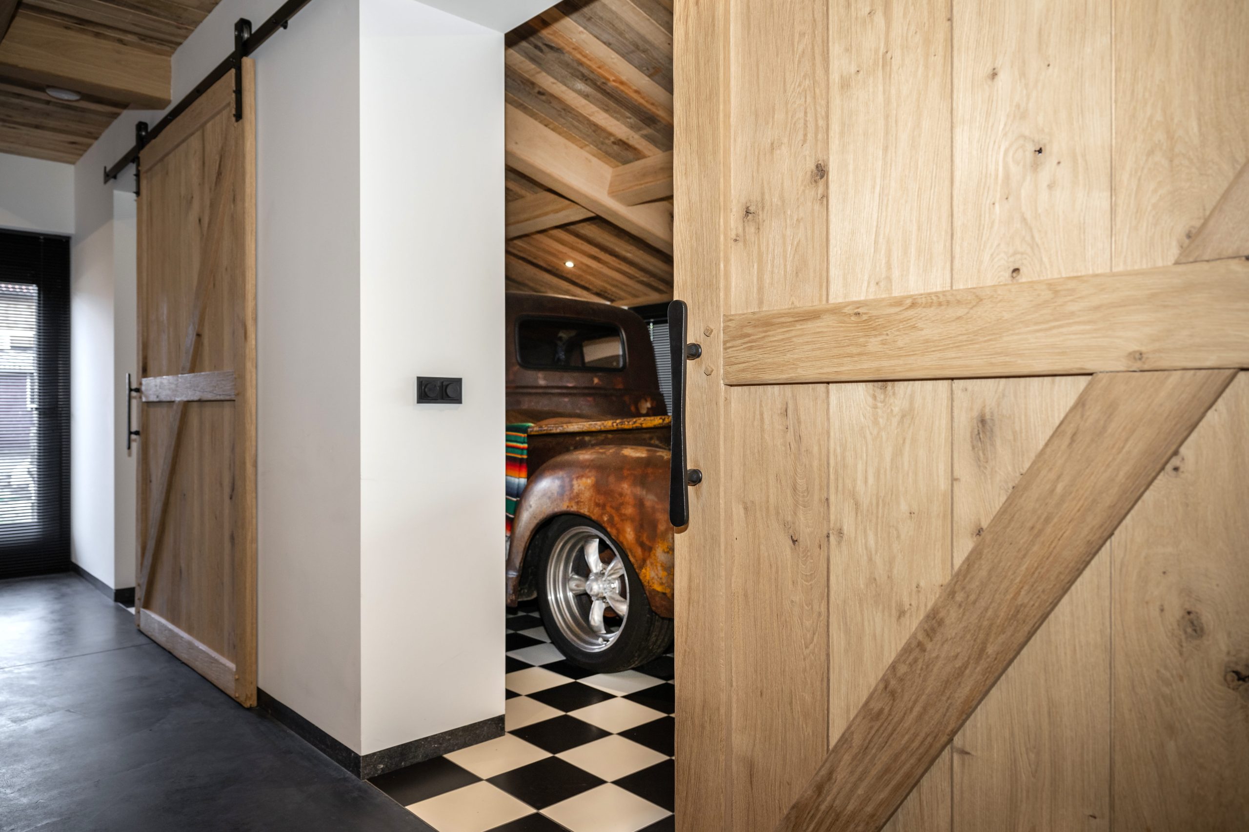 Zicht door een eikenhouten schuifdeur op een roestige vintage pick-up truck in een kamer met een zwart-wit geblokte vloer.