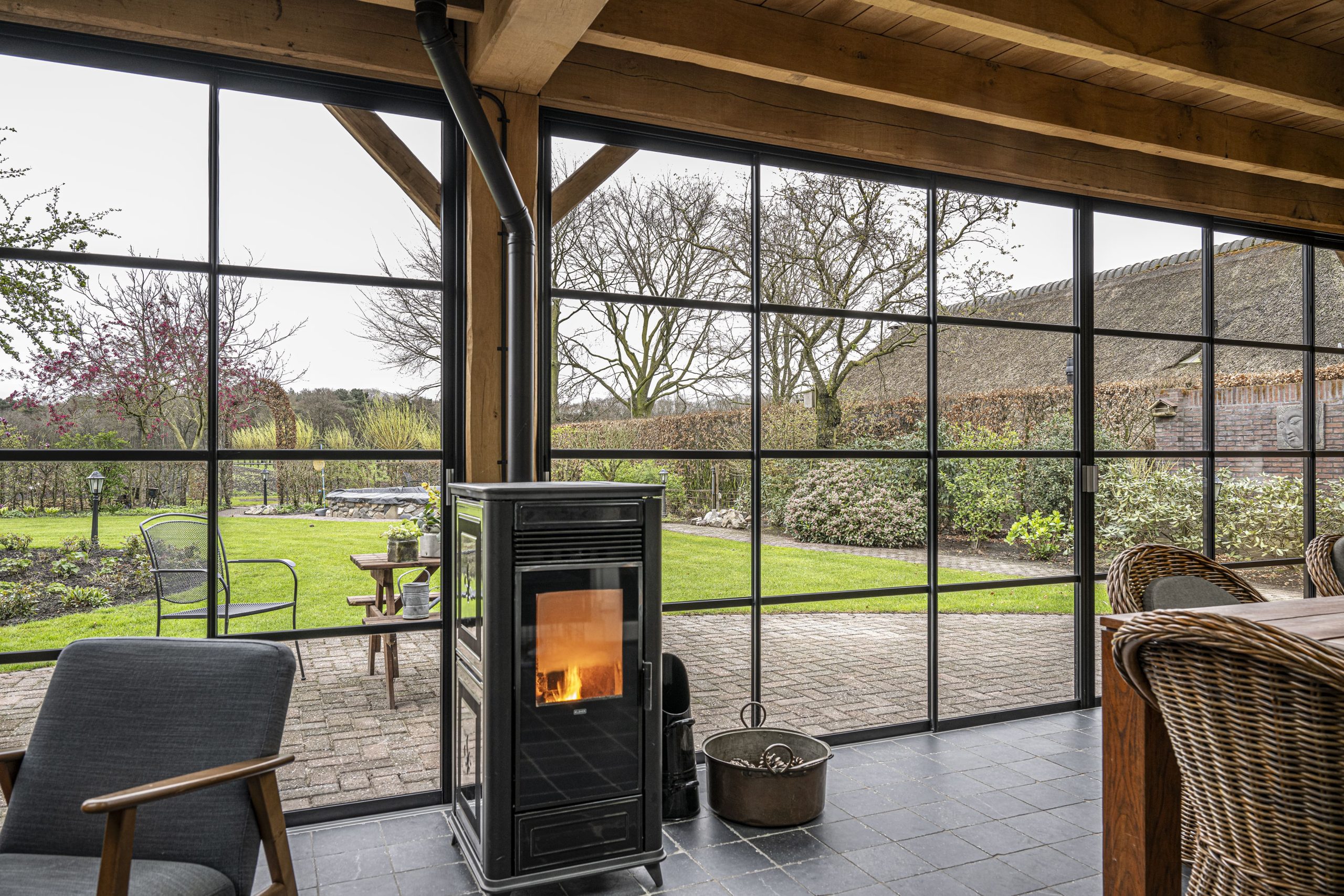 Gezellig interieur van de glazen tuinkamer met een brandende pelletkachel, een comfortabele fauteuil en weids uitzicht over de groene tuin.