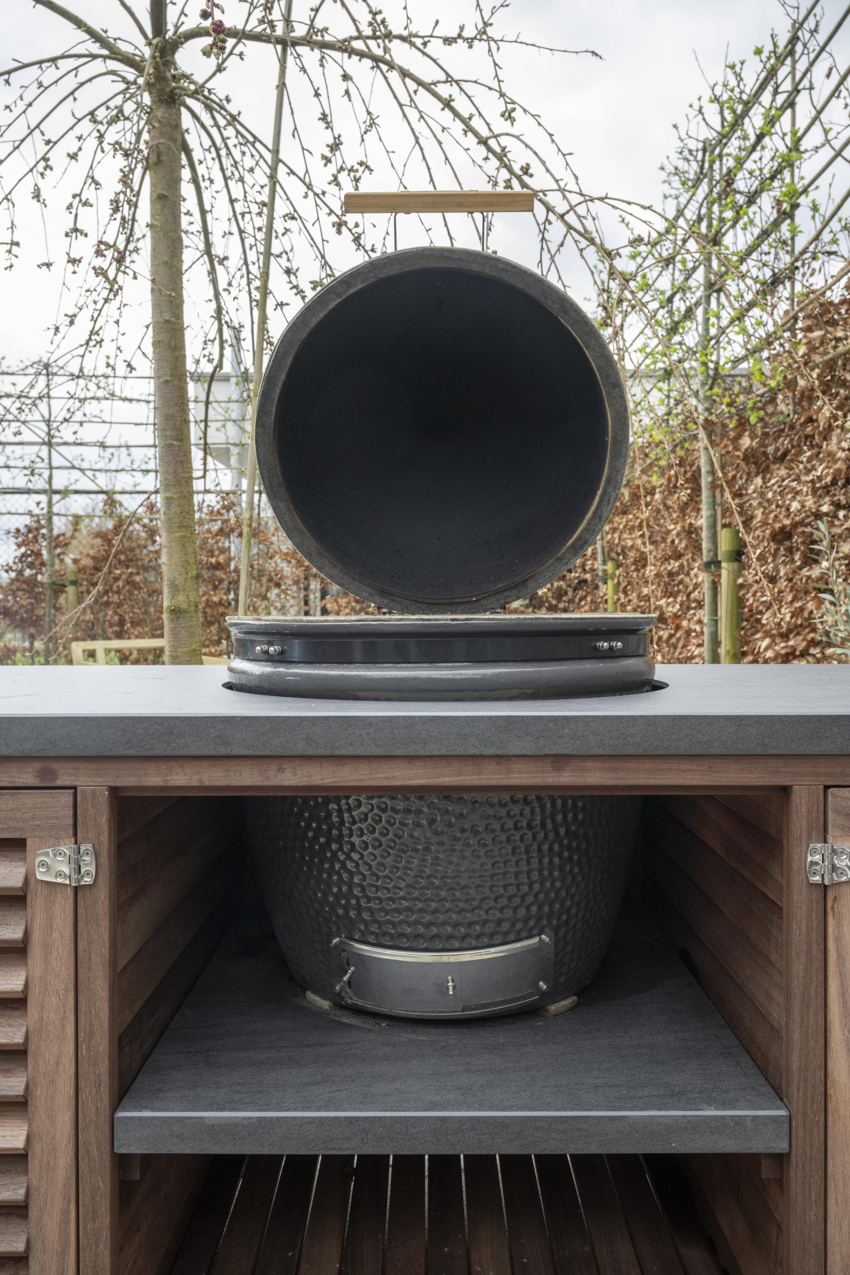 Detailopname van een geopende zwarte kamado barbecue ingebouwd in een padoek houten buitenmeubel.