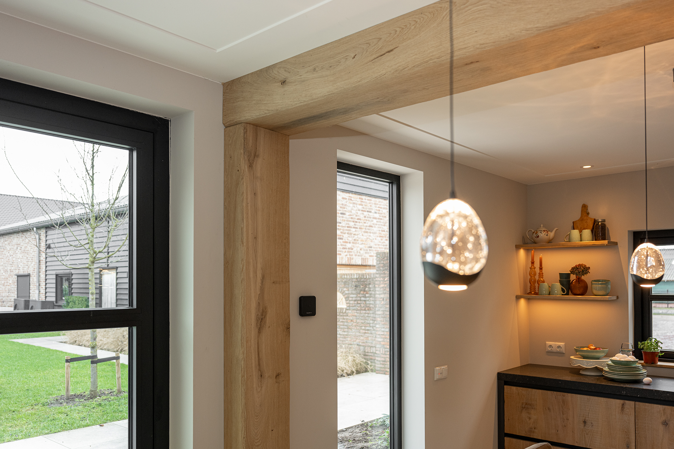 Zicht op een eikenhouten kolom naast een groot raam met uitzicht op de tuin, met een glimp van de keuken en sfeerverlichting.