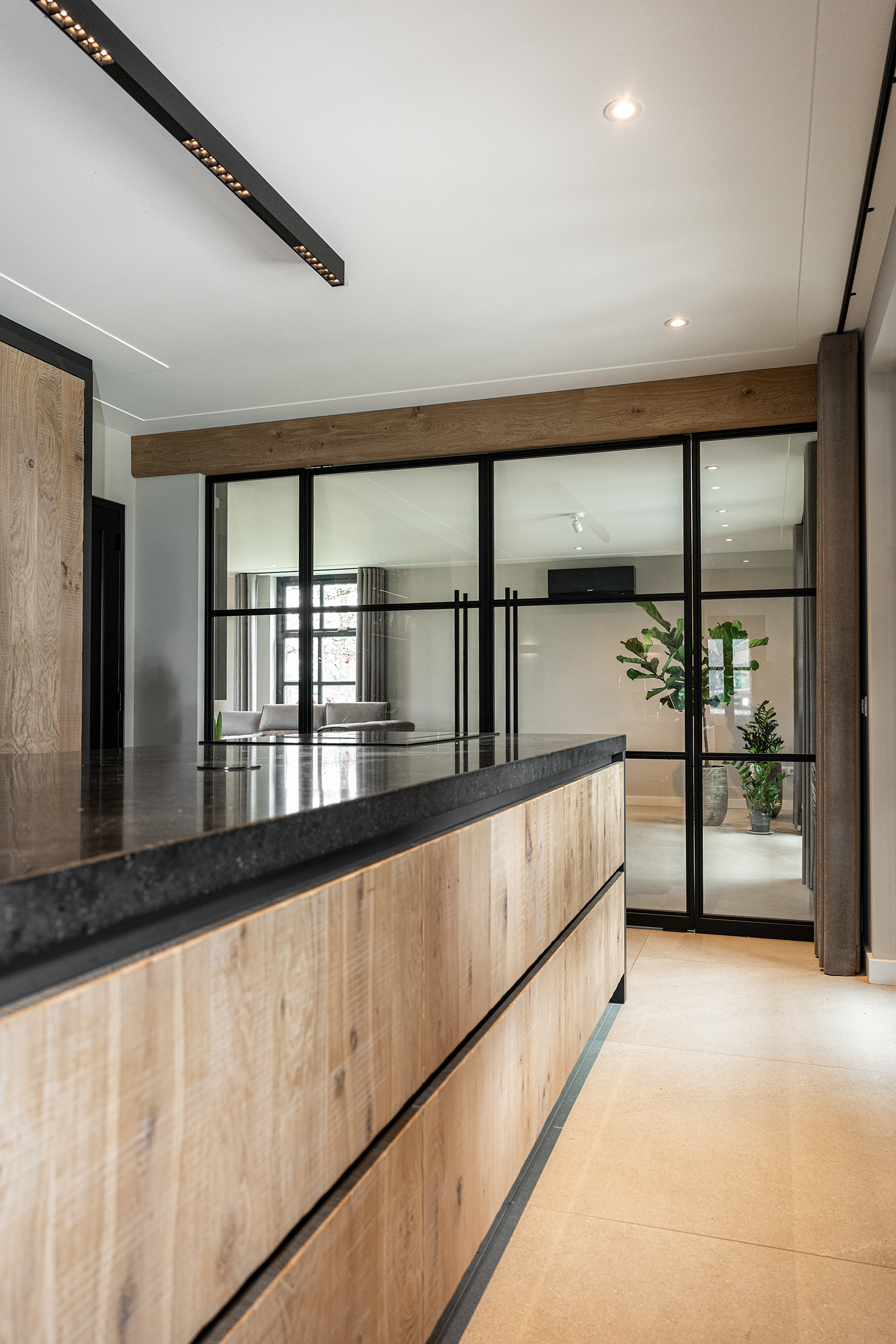 Detail van een landelijk keukeneiland met eikenhouten fronten en een dik zwart werkblad, met op de achtergrond een glazen pui met zwarte profielen.