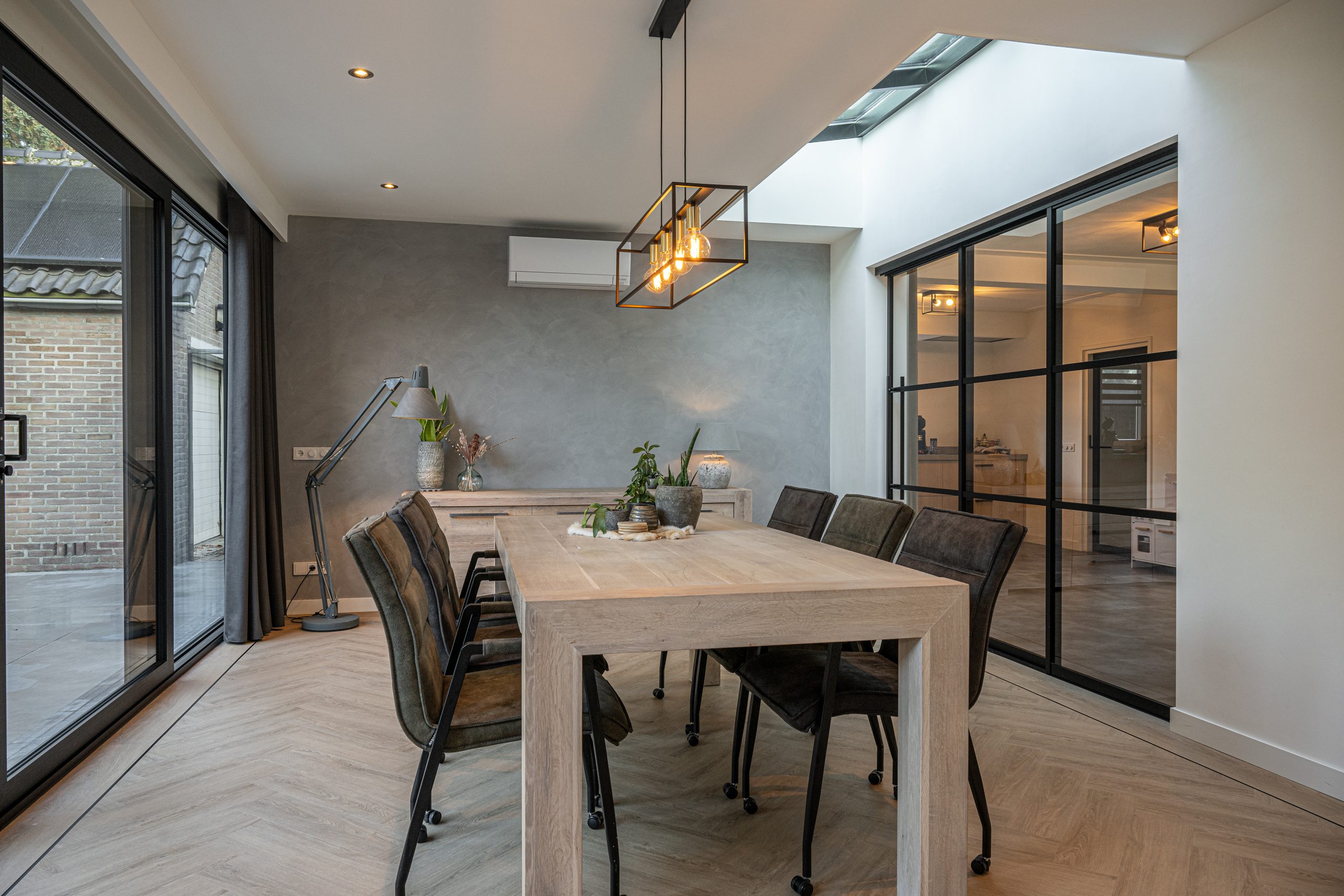 Ruime eetkamer met een houten visgraatvloer, zwarte schuifpui naar de tuin en een grote eettafel.