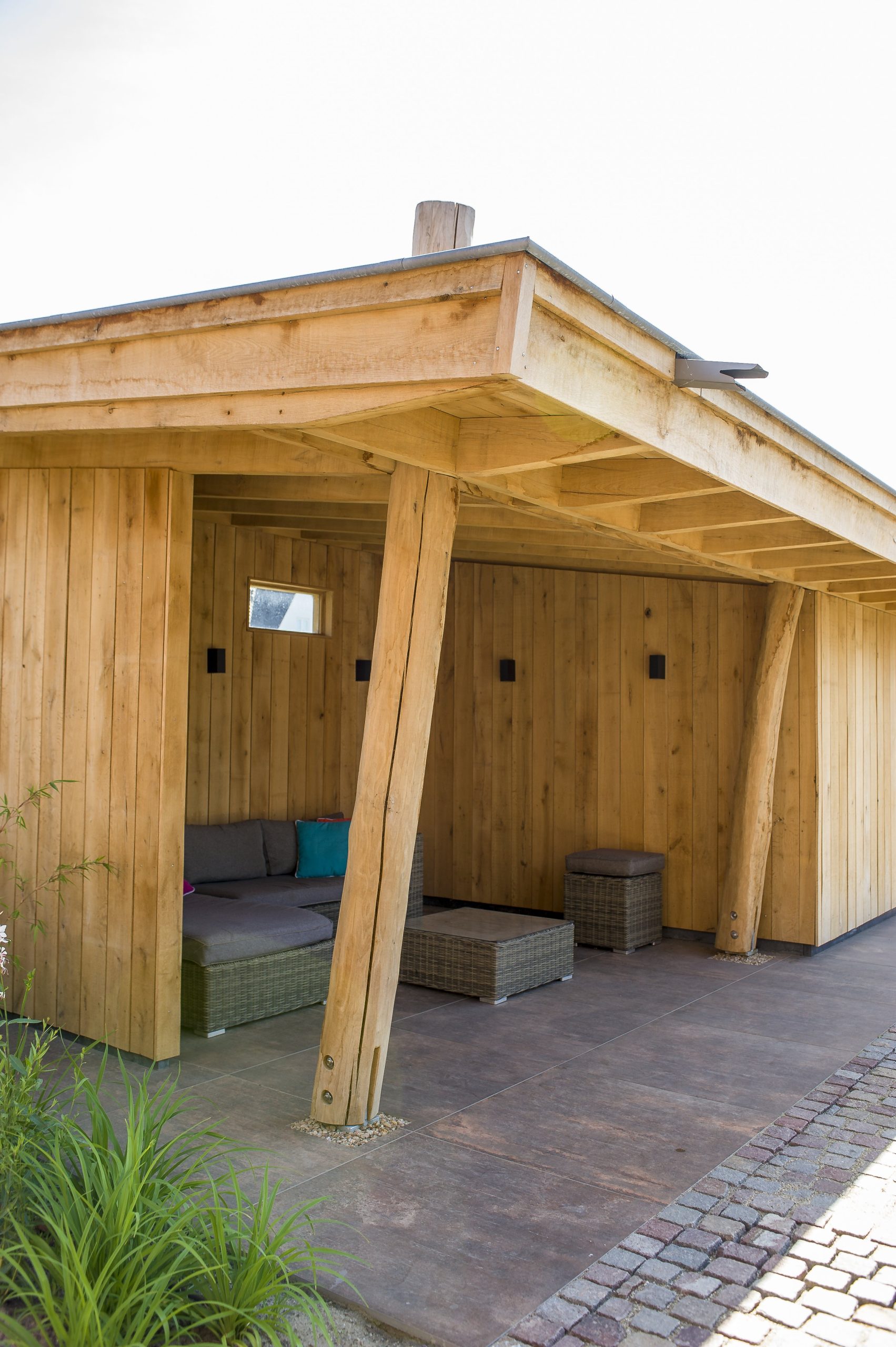 Gezellige loungehoek onder een houten overkapping met een grijze wicker hoekbank, kleurrijke kussens en een houten achterwand met kleine raampjes.
