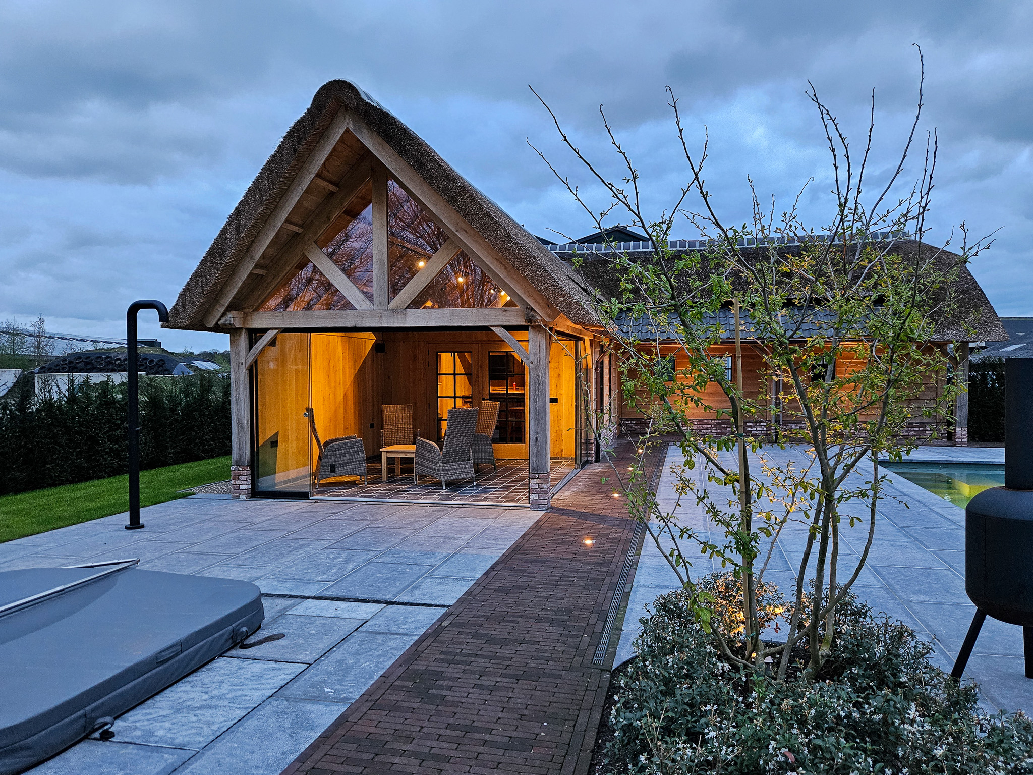 Verlicht eikenhouten tuinhuis met rieten dak en glazen schuifwand bij een terras met zwembad tijdens de schemering.