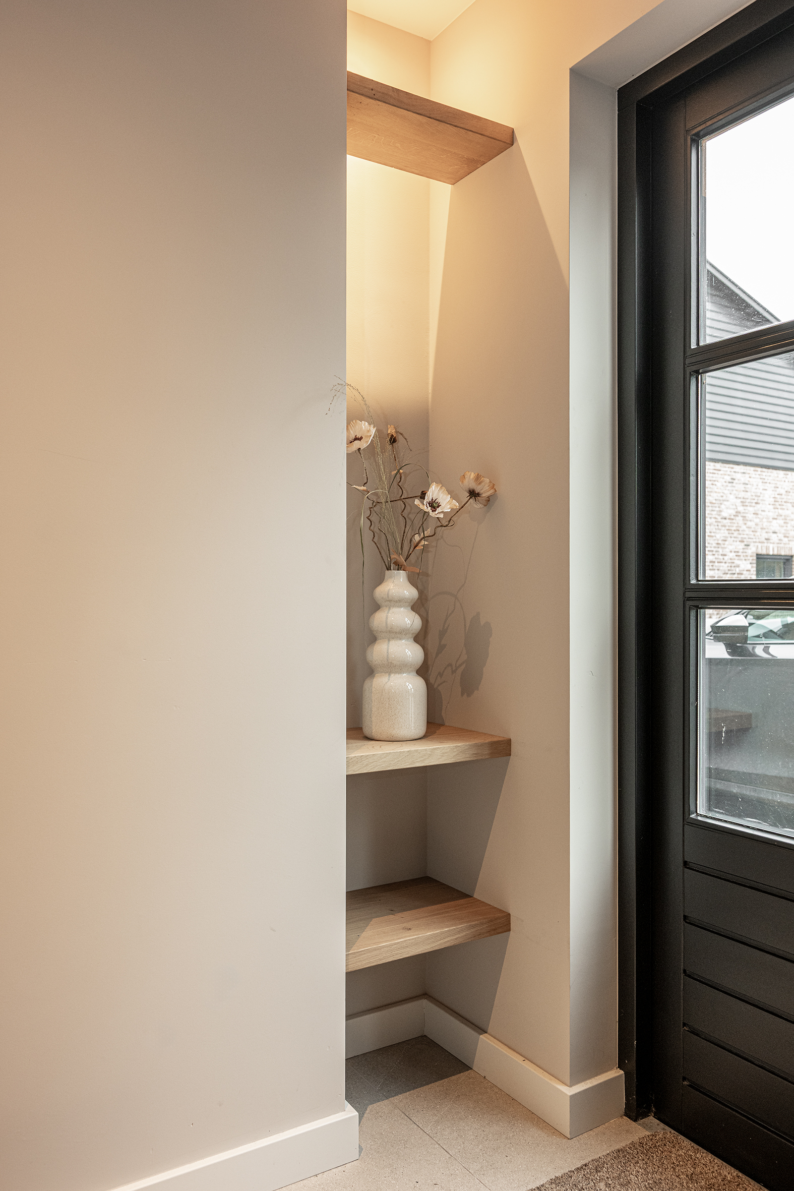 Een verlichte wandnis met maatwerk eikenhouten legplanken en een decoratieve witte vaas naast een zwarte deur.