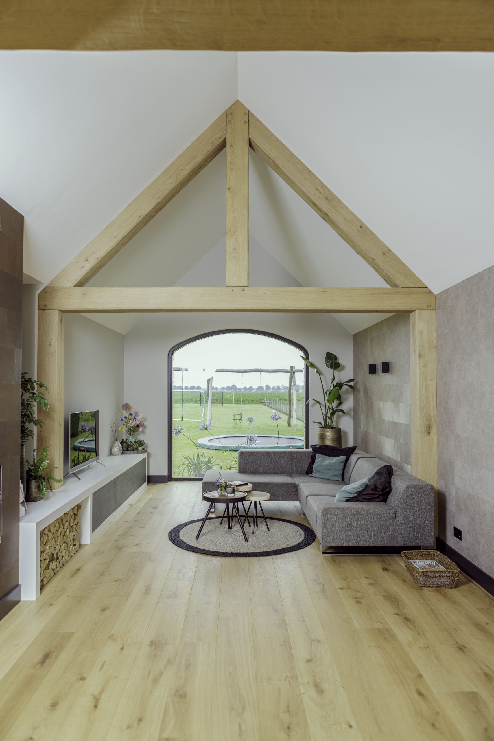 Ruimtelijke woonkamer met een indrukwekkend eikenhouten spant, grijze hoekbank en uitzicht op een grote tuin.